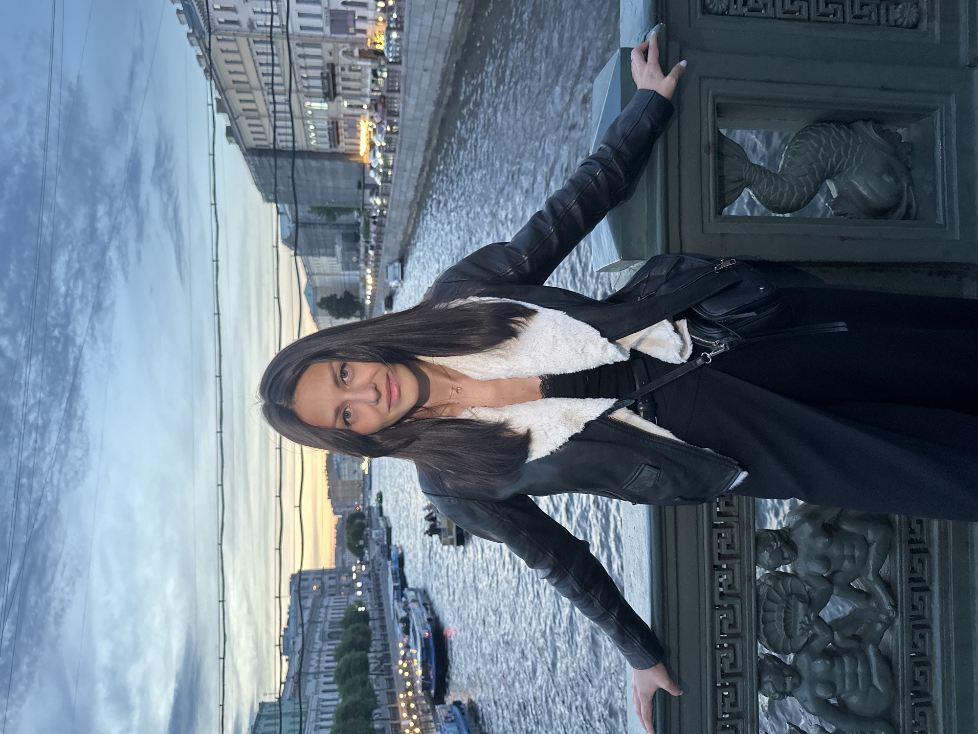 A woman stands on a bridge in Saint Petersburg, Russia, leaning on the railing. She is wearing a black leather jacket with a white fur lining, a black top, and black pants. A black crossbody bag is slung over her shoulder. The bridge railing is ornate, featuring sculpted figures of cherubs and fish. In the background, a canal runs through the city, with boats on the water and buildings lining the banks. The sky is overcast with a hint of sunset colors. The overall scene suggests an evening stroll along one of the city's many canals.
