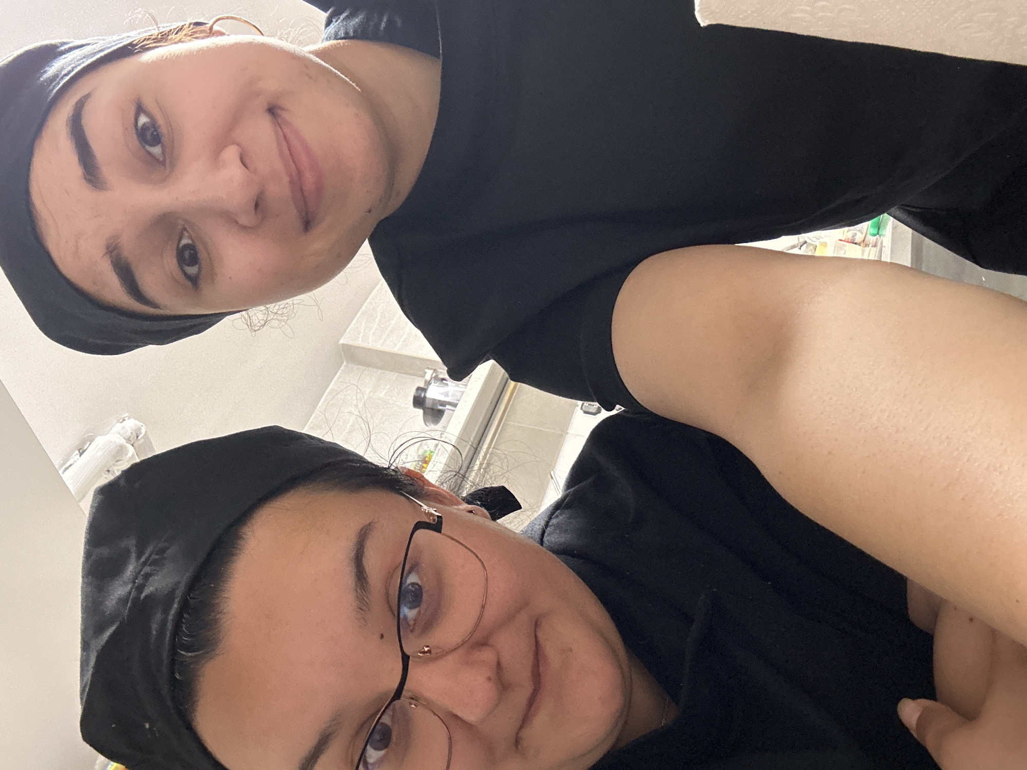 Two women are positioned close to each other in an indoor setting, possibly a kitchen or similar work area. Both are wearing black caps, possibly hairnets, and appear to be dressed in similar dark-colored attire. The woman in the foreground is wearing glasses. The camera angle is skewed, looking down on the subjects, and suggests the photo was taken in close proximity. The background is slightly blurred, but reveals what looks like kitchen fixtures and possibly some packaged goods on shelves. There is no readily discernible information in the image to place it specifically in Burgas, Bulgaria.
