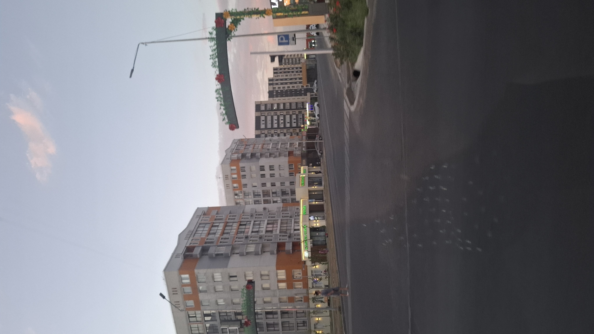 The image depicts a street scene in Shymkent, Kazakhstan, likely during dusk. In the background, several modern apartment buildings are visible. The building on the left is tall and features an orange and gray facade with numerous windows. To its right, a row of shorter, multi-story buildings line the street, many with illuminated storefronts at ground level. A street lamp stands prominently in the mid-right of the image, topped with a decorative arch adorned with artificial flowers and foliage. A "P" parking sign is also visible near the street lamp. The street itself is paved, with a crosswalk in the foreground. A single person in a gray shirt and shorts is walking on the sidewalk to the left of the apartment buildings. Several cars are parked or driving on the street. The sky is a pale blue with a hint of pink from a distant cloud. The foreground of the image is dominated by the dark surface of the road, reflecting some light in a scattered pattern.