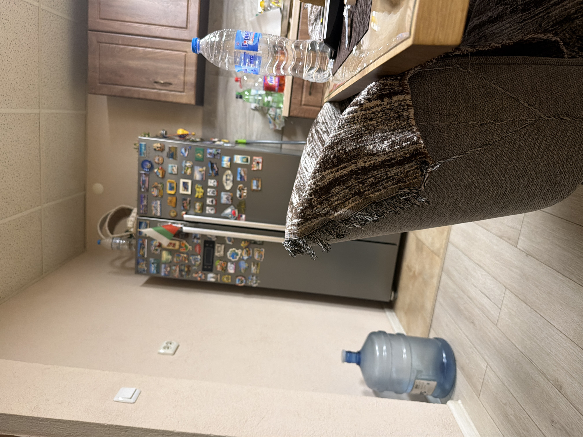 The image shows a kitchen interior. A refrigerator covered in magnets is visible against a wall with light switches. Two large water bottles, one partially obscured and the other in the foreground, are present. A table with a patterned rug hanging over its edge is in the upper right corner. A smaller bottle of water is near the top of the image, sitting on the table. The location is Panagyurishte, Bulgaria.
