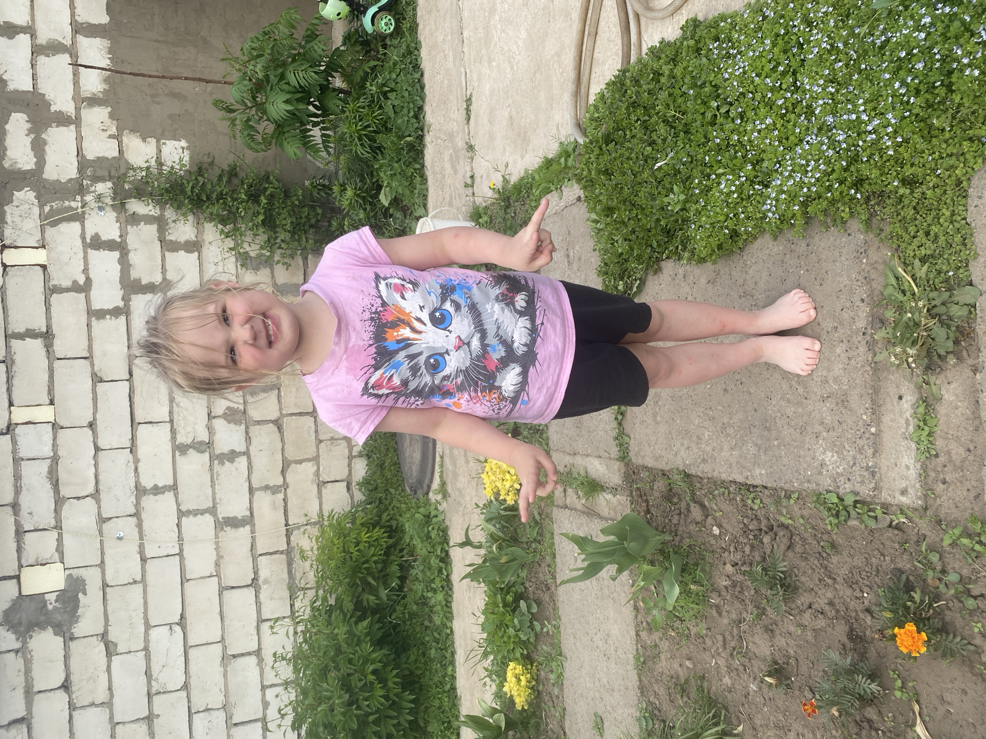 A young girl stands barefoot on a concrete path surrounded by greenery, with a brick wall in the background. She wears a pink shirt with a colorful cat graphic and black shorts. Her blonde hair is messy, and she smiles while holding up one finger on her right hand. The environment includes various plants, including ground cover with small blue flowers, as well as some yellow and orange flowers. A hose and a child's scooter are visible in the background. The location is identified as Chaikovsky, Russia.