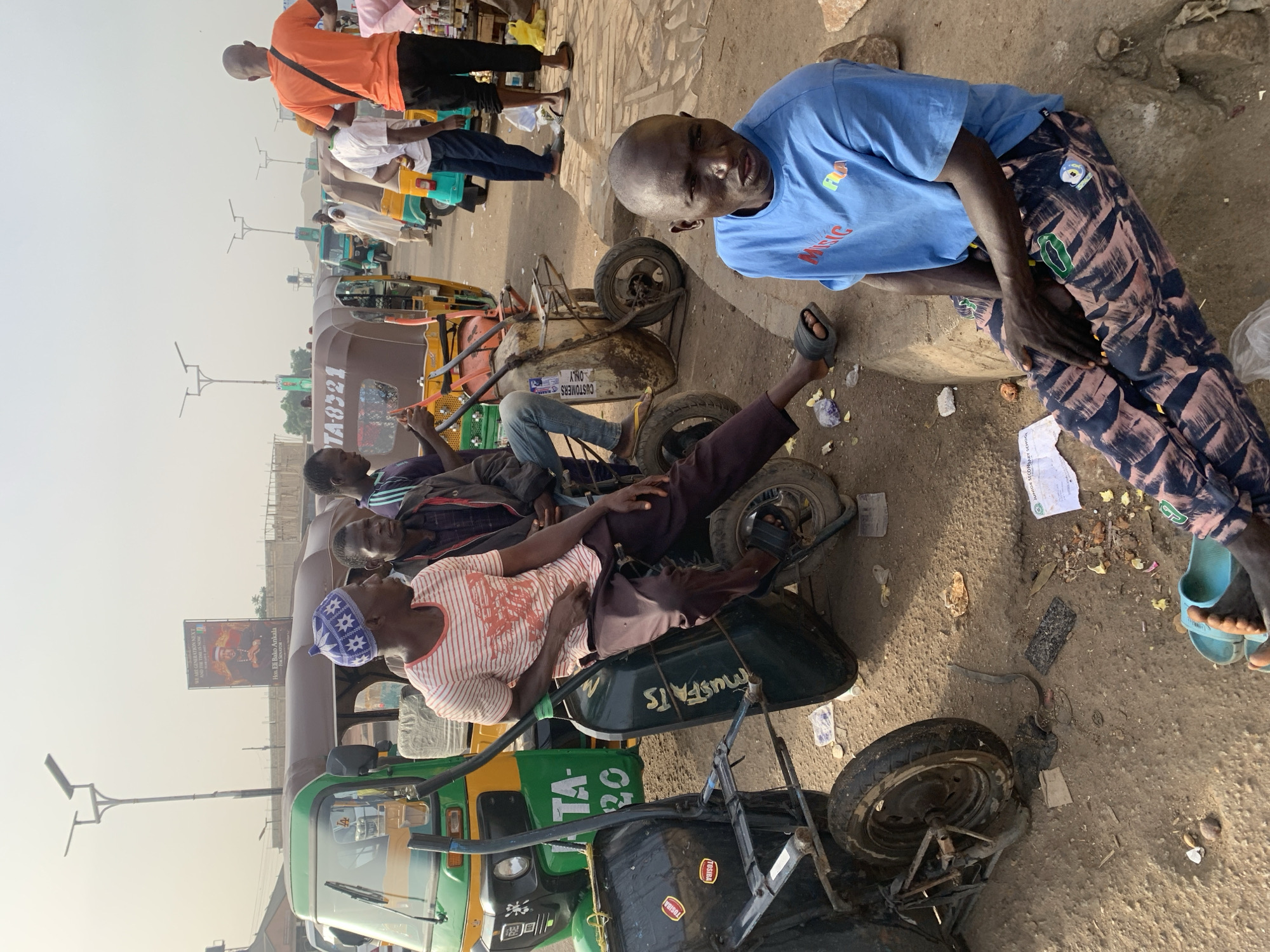 The image shows a street scene in Jos, Nigeria. There are several auto rickshaws (tuk-tuks) parked along the side of the road, with men either resting or sitting on them. One man is lying down on a tuk-tuk. Another man in a blue shirt is looking directly at the camera. A man in an orange shirt is standing near a small kiosk. The road appears to be paved, with some dirt and debris on the side. A banner advertisement is visible on one of the tuk-tuks. Text is visible on the tuk-tuks and shirts, including "TOSIBA", "TA-20", "MUSFATS", "TA-8321", and "MISIC".