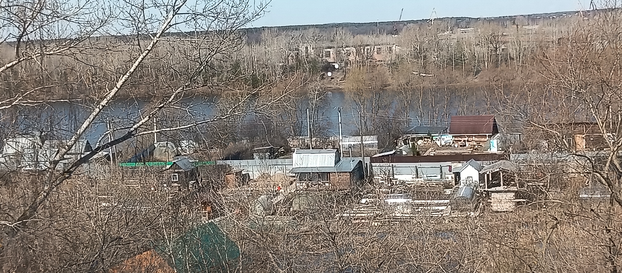 The image shows a view of a neighborhood in the city of Unknown, Unknown. The foreground consists of bare trees with no leaves. Behind the trees is a collection of small houses and buildings with various roof types and construction materials, including wood and metal. A river or body of water is visible behind the houses, with trees and some buildings visible on the opposite bank. There is smoke rising into the air in the middle ground of the image. The sky is bright and clear.