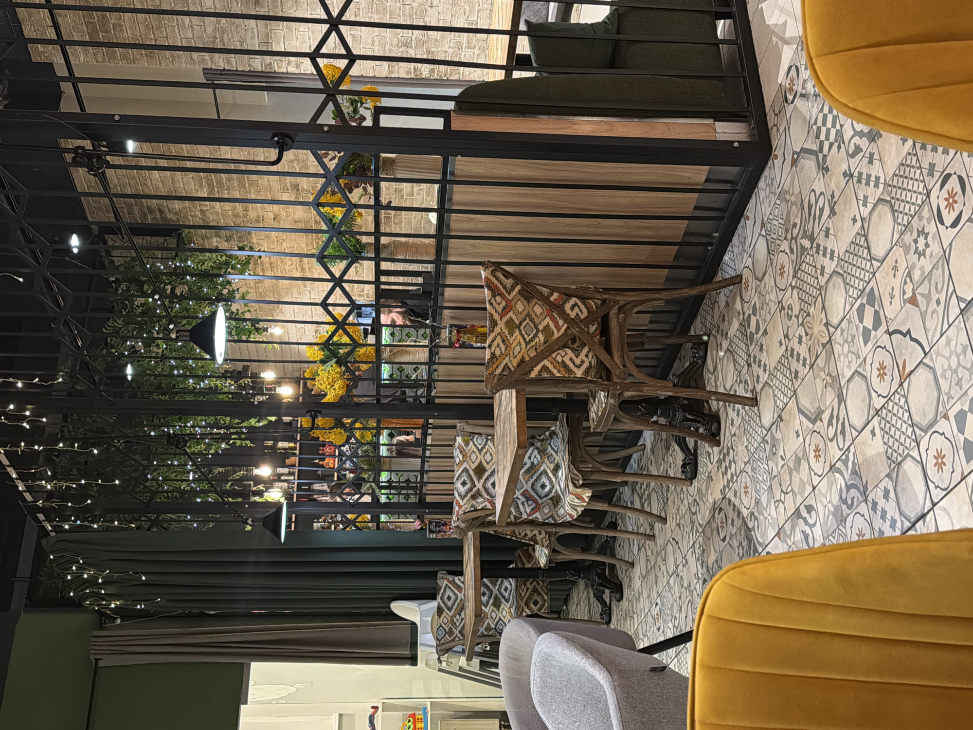 The image shows an interior scene that appears to be a cafe or restaurant, likely in Izhevsk, Russia. The foreground features a few tables and chairs; the chairs have patterned upholstery. Some chairs are modern with gray fabric, and a yellow upholstered chair is partially visible on the right. The floor is covered in patterned tiles. Behind the seating area is a decorative iron or metal structure that stretches across the width of the frame, with wooden shelves and potted plants. Through the structure, one can see details of another area. There are warm lighting fixtures and greenery, suggesting a deliberate design aesthetic. A person is visible beyond the metal structure. The overall atmosphere is decorative and intimate.