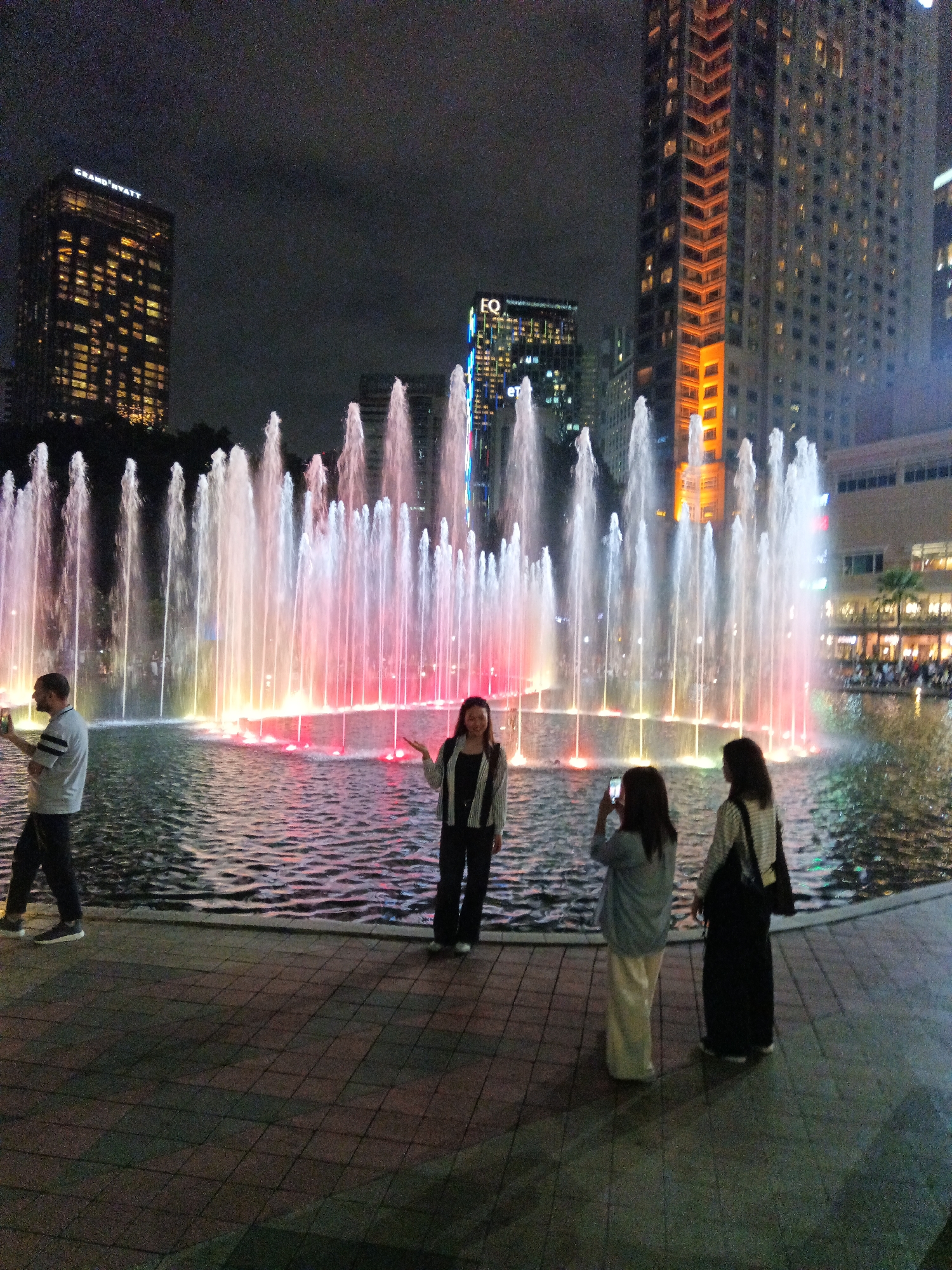 The image depicts a night scene at the Symphony Lake at KLCC Park in Kuala Lumpur, Malaysia, characterized by a large, illuminated fountain. The fountain features multiple jets of water, some reaching considerable heights, illuminated with vibrant pink, red, and yellow lights. The water reflects the colorful lights, creating a dynamic visual effect.
In the foreground, four individuals are present. On the left, a man wearing a grey t-shirt and dark pants is standing and holding a mobile phone. To his right, a woman in a black and white jacket and black pants is posing with her arms outstretched. Further to the right, two women are visible. One, in a light blue cardigan and cream-colored pants, is taking a photograph with her phone, aiming it towards the fountain. Beside her, another woman in a striped top and black skirt stands with her back to the camera, carrying a bag over her shoulder.
In the background, the iconic Petronas Twin Towers are partially visible, along with other illuminated high-rise buildings. The sky is dark, indicating nighttime. The ground in the foreground is paved with square tiles. The overall atmosphere is one of a tourist attraction at night, with people enjoying the fountain display and the surrounding city lights.