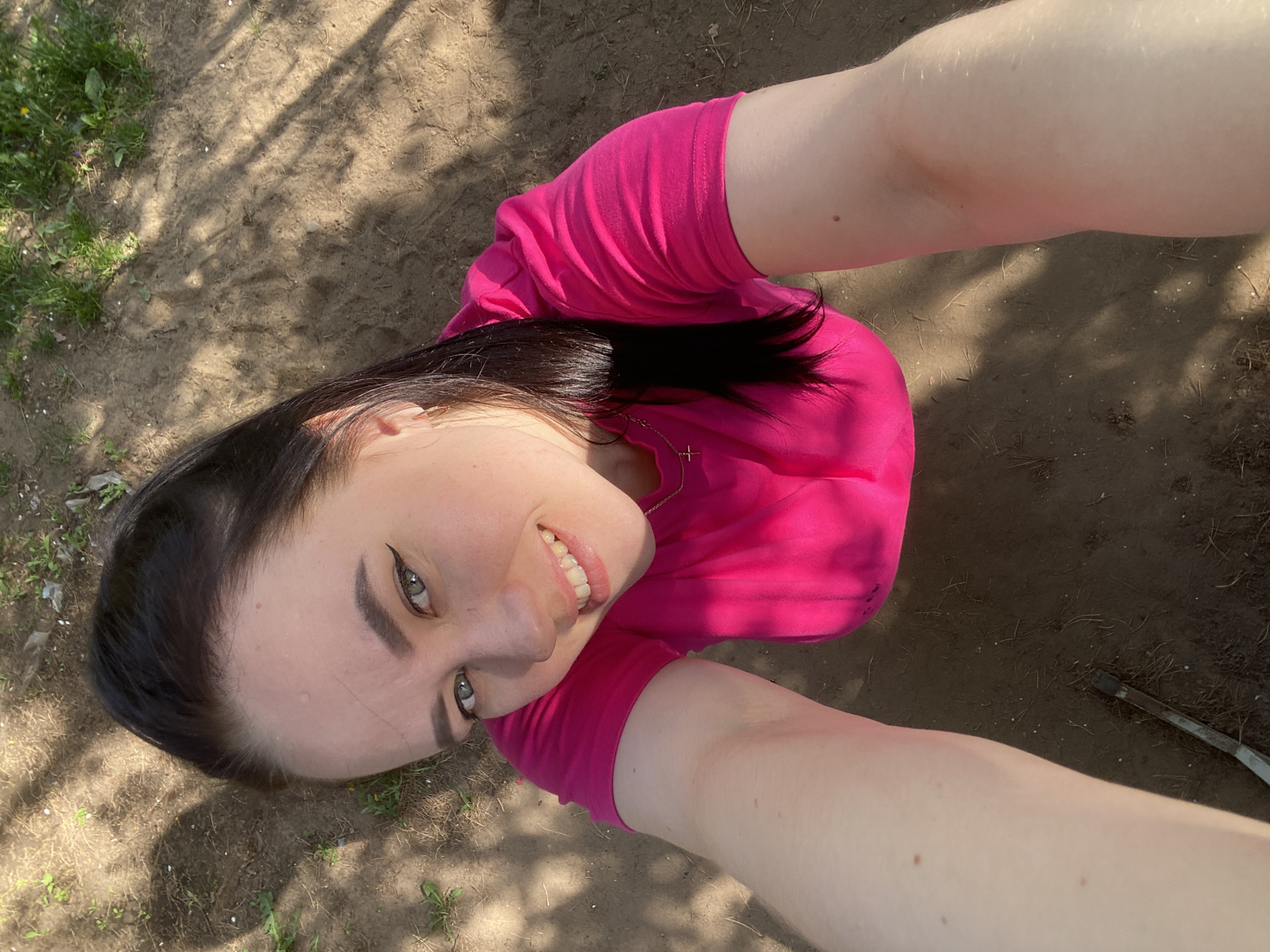 A high-angle selfie shows a woman in a bright pink shirt standing on dirt-covered ground under dappled sunlight. The ground also contains a small amount of green vegetation, and the woman's arms are visible, holding the camera above her head. The woman appears to have dark hair and light skin. The precise location in Chaikovsky, Russia, cannot be determined from the image alone.