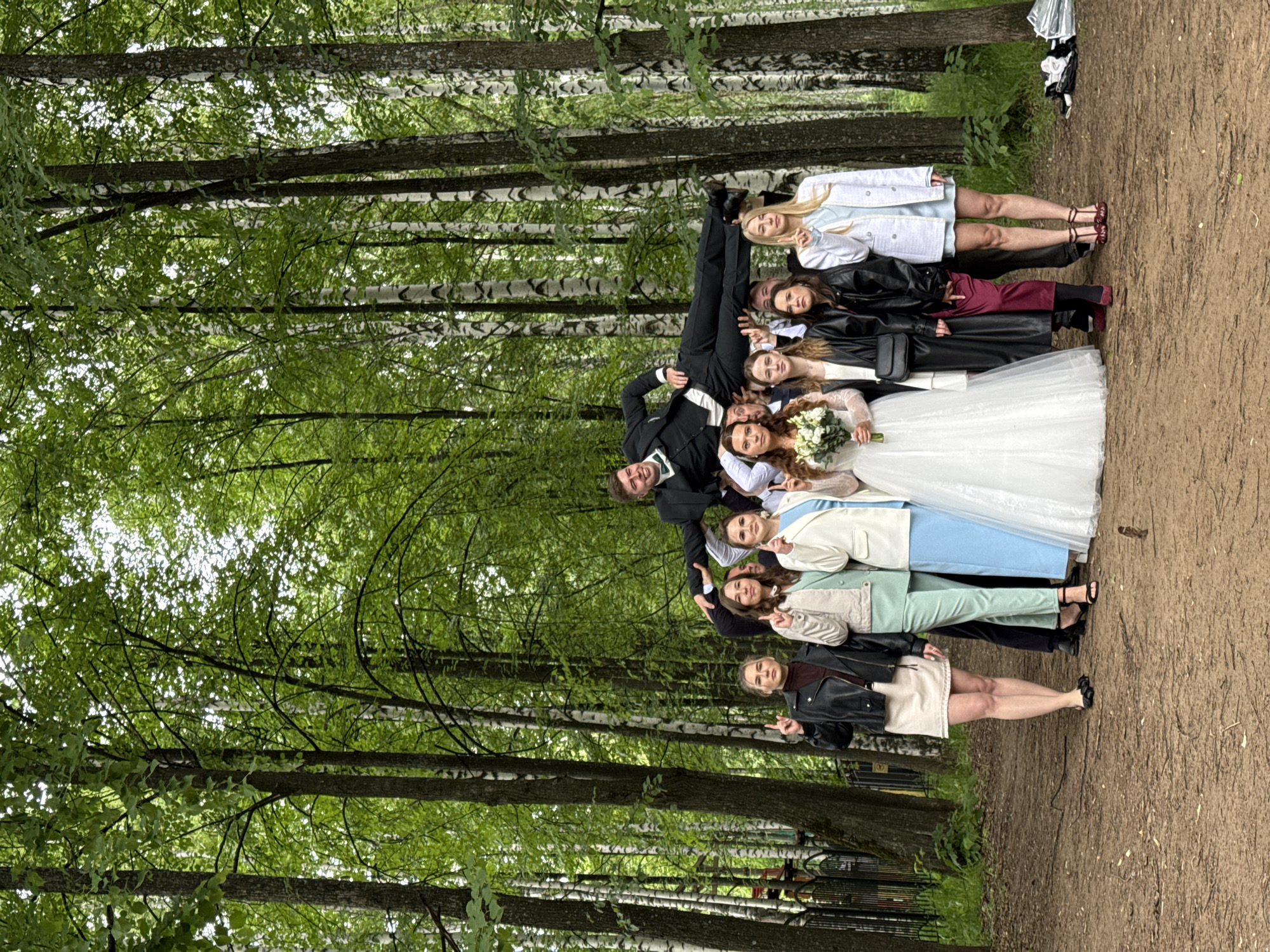 The image depicts a lively group of approximately 10-12 young adults, likely a wedding party, posing for a photograph in a birch forest in Chaikovsky, Russia. The setting suggests a pleasant spring or summer day, with abundant green leaves on the tall, slender birch trees that dominate the background and canopy.
At the heart of the group, a man dressed in a black suit, white shirt, and bow tie (presumably the groom) is playfully held upside down by his legs by another man, eliciting smiles and laughter from those around him. Standing next to this playful scene is a woman in a traditional white wedding dress, holding a bouquet of white flowers, who is most likely the bride. She is smiling broadly, contributing to the joyful atmosphere.
The other individuals in the group are dressed in a variety of semi-formal and smart-casual attire, including suits, light-colored blazers, leather jackets, dresses, and skirts. Many are looking towards the camera with happy expressions, some making hand gestures. The ground beneath them is a light brown dirt or gravel path. In the distance, beyond the immediate group, more birch trees create a dense, natural backdrop. A small pile of white and black fabric, possibly an umbrella or discarded item, is visible on the ground to the right. The overall impression is one of celebration and camaraderie in a picturesque natural environment.