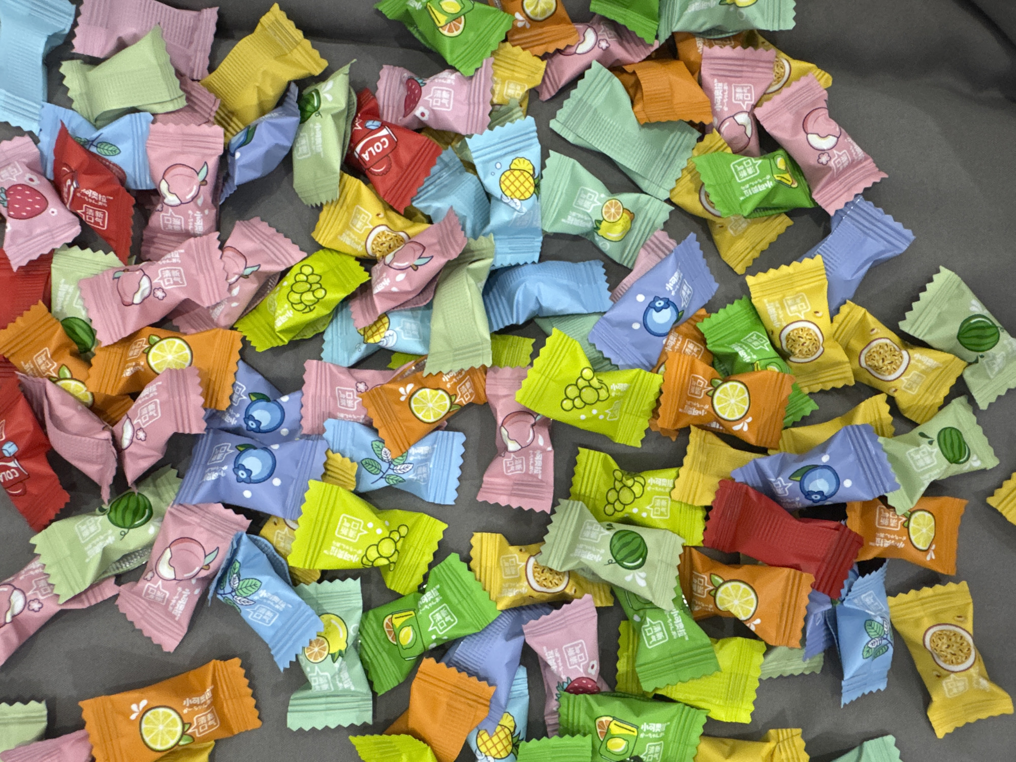The image shows a collection of individually wrapped candies scattered on a gray surface. The wrappers come in various colors, including pink, red, orange, yellow, green, and blue. Each wrapper features a different fruit or flavor illustration, such as strawberry, cola, lemon, lychee, watermelon, grape, blueberry, and passion fruit. Some wrappers also include Chinese characters and brand names.