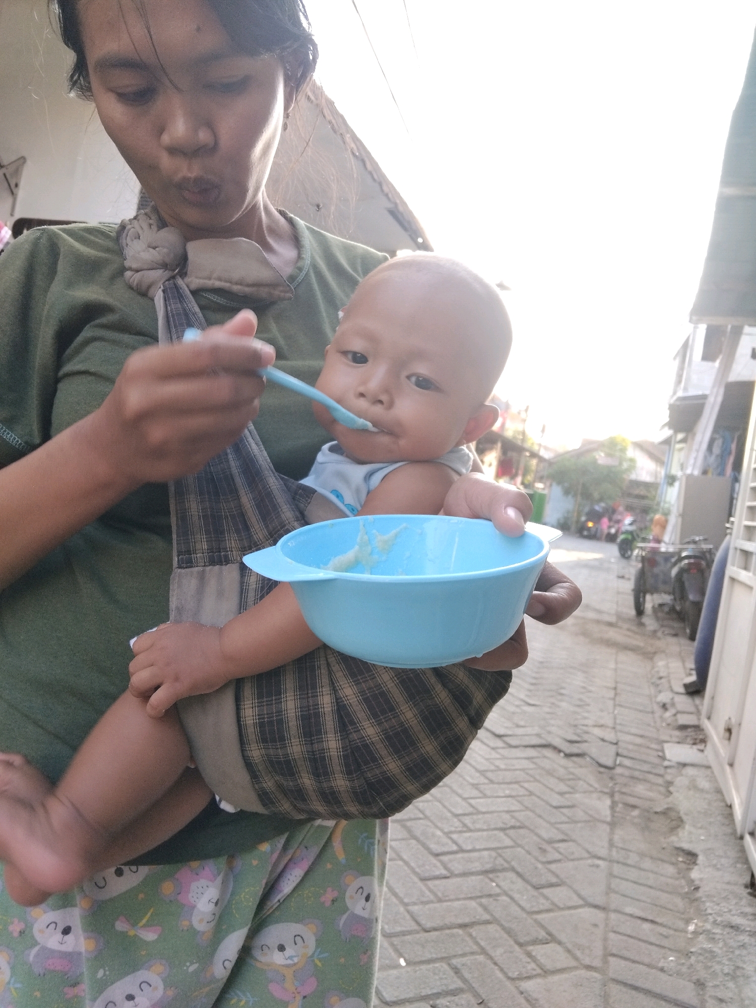 In Kepuh Kiriman, Indonesia, a person is feeding a baby from a blue bowl. The baby is in a sling carrier on the person's chest. The person is wearing a green shirt and the baby is wearing a blue shirt. The background shows a narrow street with buildings and a few motorbikes. The ground is paved with bricks. The sun is shining brightly, casting long shadows.