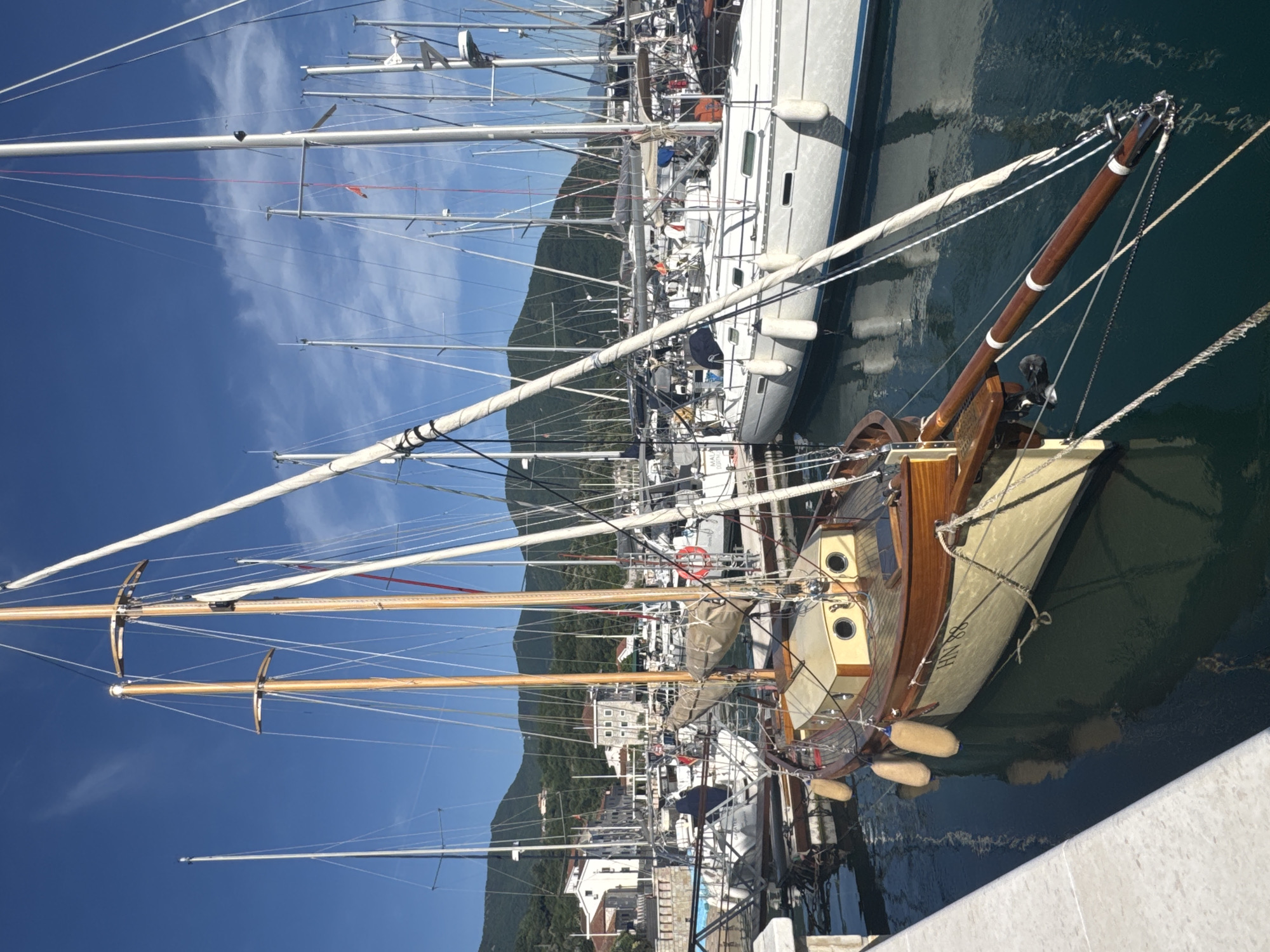 The image shows a harbor scene in Meljine, Montenegro. Several boats are docked. The predominant boat is a classic wooden sailboat, with tan hull, dark wood trim, and cylindrical fenders. It is moored alongside a concrete wall, possibly a pier or quay. A larger, modern white sailboat is also visible, docked next to the wooden one. In the background, a mountain rises, and some buildings of the town are visible at its base. The sky is blue with some wispy clouds. Many masts from other sailboats are visible, filling the sky with linear elements.