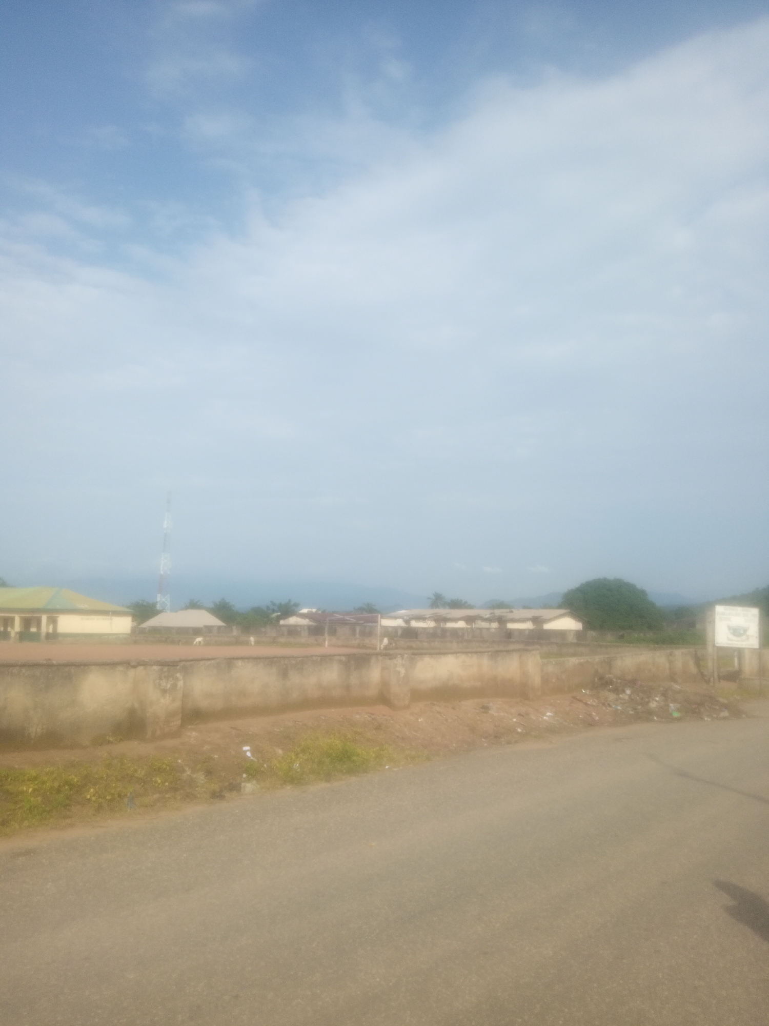 This media file captures an outdoor scene in Serti, Nigeria. The image displays a collection of buildings with light-colored walls and green or beige roofs. A tall, white and red striped radio tower stands prominently in the background, to the left of the buildings. In front of the structures, there is a low, concrete wall. Beyond the wall, the ground appears to be a mix of dirt and sparse, dry vegetation. A paved road runs along the foreground of the image. The sky above is a pale blue with scattered white clouds, suggesting it is daytime, likely morning or afternoon. The overall impression is one of a somewhat arid, possibly rural or semi-urban environment. There is a sign visible on the right side of the image, but any text on it is illegible due to the image quality. No people or specific activities are clearly depicted, though the presence of a road and buildings indicates a populated area. The weather appears to be clear.