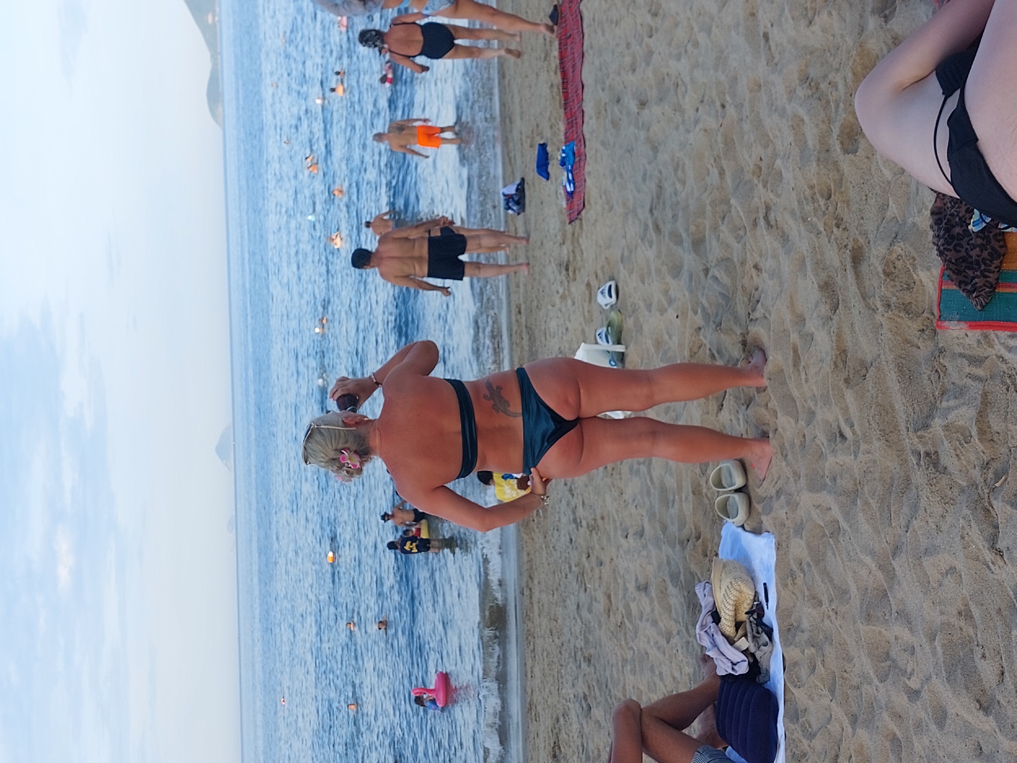 A diverse group of people are enjoying a sunny day at a beach in Nha Trang, Vietnam. The water is calm and blue, with several individuals swimming and playing in the ocean. On the sandy shore, people are relaxing, sunbathing, and interacting with each other. Objects scattered on the beach include towels, bags, and footwear. The overall scene depicts a lively and casual atmosphere at a popular tourist destination.