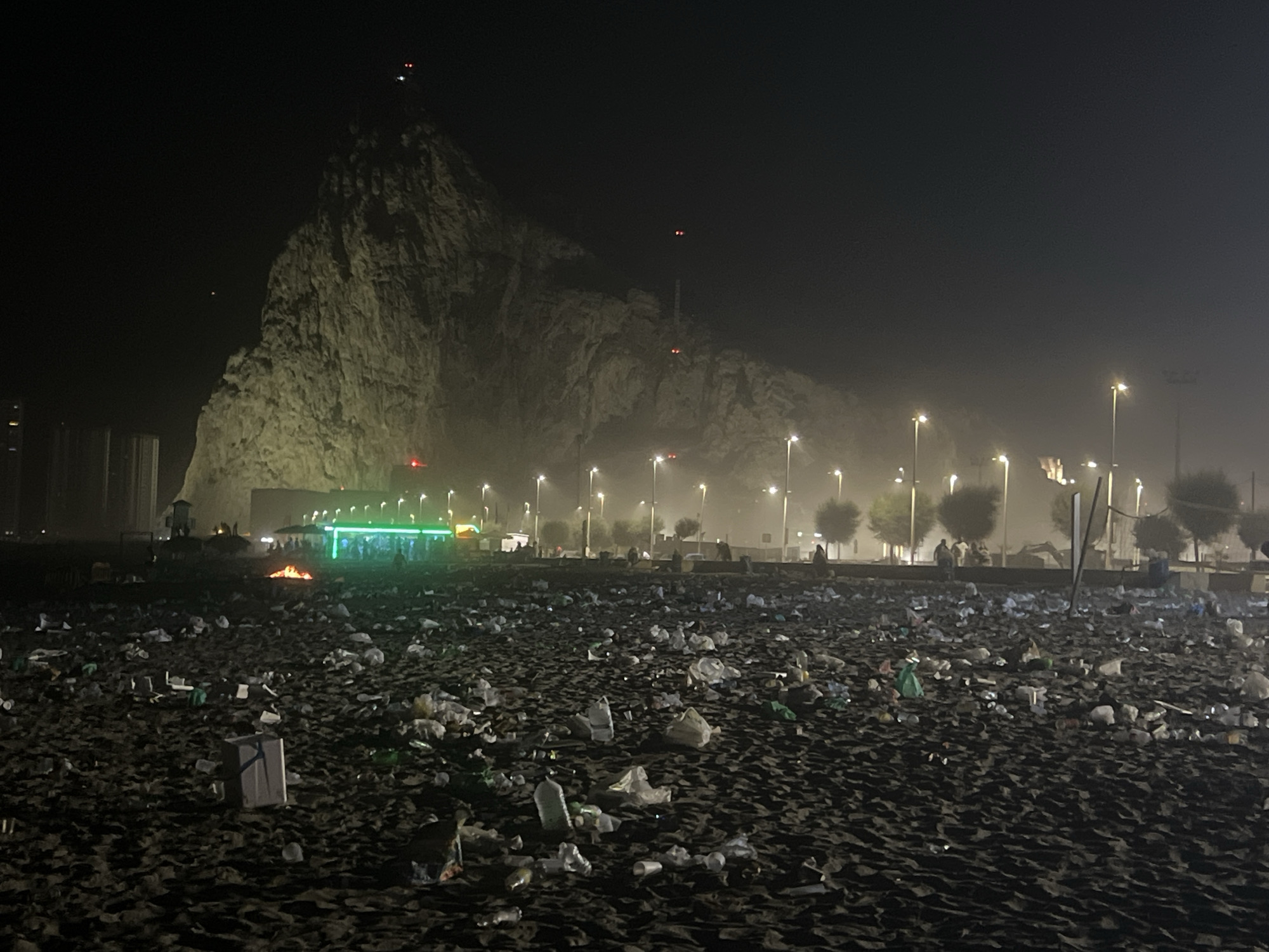 The image depicts a beach at night, heavily littered with plastic bottles, bags, and other debris. In the background, a large, rocky cliff face looms, illuminated by artificial lights. Several streetlights line the edge of the beach, casting a hazy glow on the scene. A building with green lights is visible in the middle ground, with a small bonfire burning in front of it. Silhouetted figures of people can be seen near the building and further down the beach. The sky is dark, suggesting nighttime, and a light fog or haze is present, contributing to the diffused lighting. The overall impression is one of neglect and environmental concern, with the natural landscape overshadowed by human-generated waste. There are no visible signs or text in the image. The location is identified as La Línea de la Concepción, Spain.