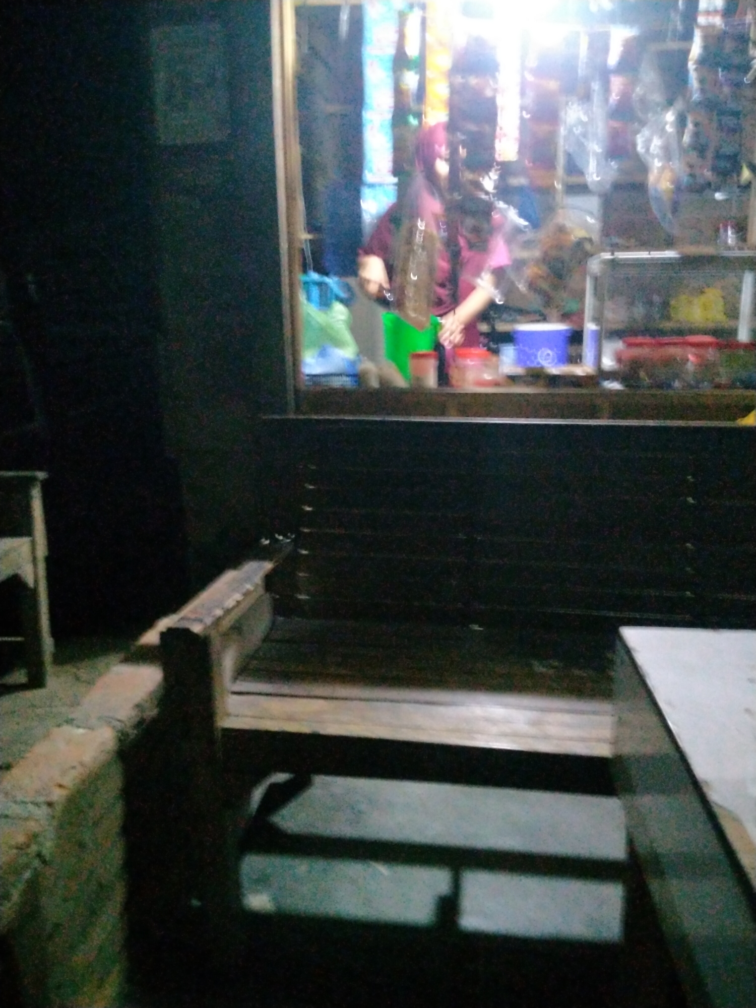 The image captures a dimly lit scene within a small shop or stall in Kruengbaroblangme, Indonesia. A person wearing a maroon headscarf and a pink top is partially visible behind a counter, their hands engaged in an unseen task. In front of the counter, a dark wooden bench is prominently featured, with a brick-like structure to the left and a white tabletop to the right. The background shows shelves filled with various items, suggesting a retail environment. The overall atmosphere is indistinct due to low light and motion blur.