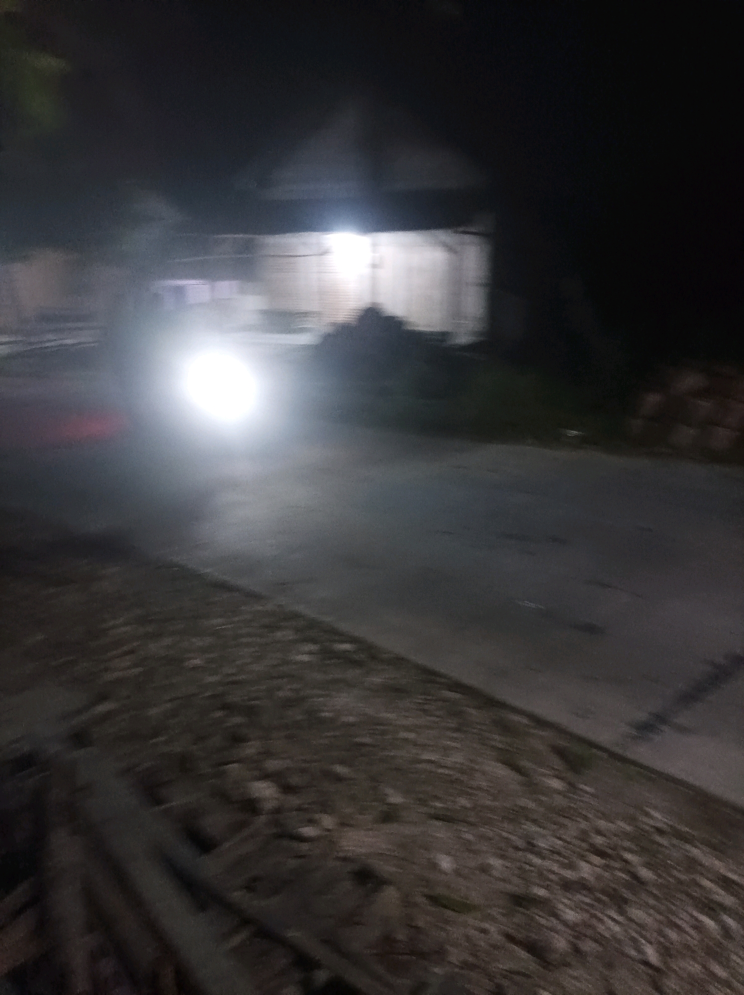 In Canga'an, Indonesia, at night, a car with its headlights on drives on a paved road. The headlights are very bright and create a glare. The road is on the right side of the image, with a dirt shoulder on the left. In the background, there is a building with lights on. The image is blurry, suggesting motion.