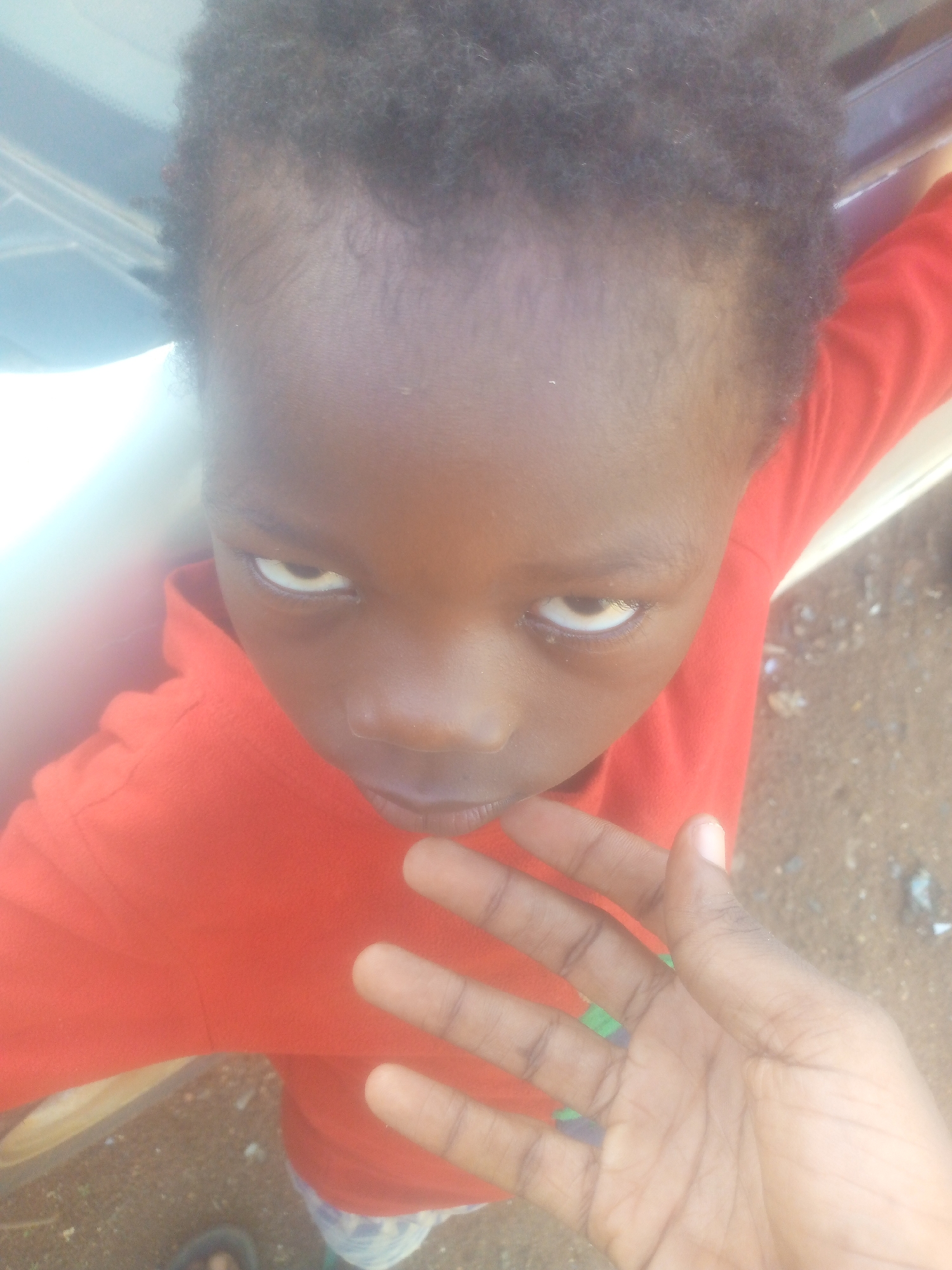 A close-up, high-angle shot captures a young child with dark, curly hair and dark skin. The child is wearing a bright red long-sleeved shirt. Their face is angled slightly upwards, directly looking into the camera with their eyes partially rolled upwards, revealing more of the whites of their eyes. Their lips are slightly parted.
In the foreground, a hand, possibly belonging to the child or another individual, is extended towards the camera, with fingers slightly splayed and the palm visible. The background consists of a light-colored, somewhat blurry object or structure on the left, which might be part of a vehicle or building, and a brown, textured ground on the right, suggesting an outdoor setting. The lighting is bright, indicating it is daytime. No text is visible in the image.