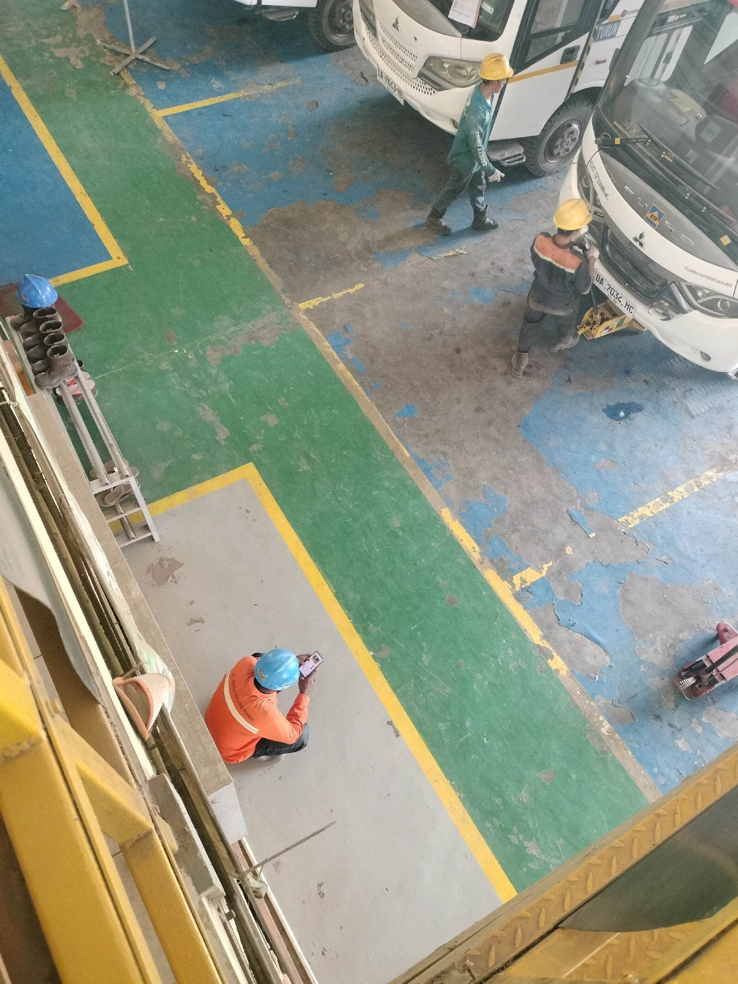 In a dimly lit indoor bus maintenance facility, possibly in Maburai, Indonesia, two men in work attire are engaged in activity. One man, wearing a blue helmet and an orange long-sleeved shirt, is kneeling on the floor while looking at his phone. Another man, wearing a yellow helmet and a blue long-sleeved shirt with dark pants, is standing and appears to be working on a white bus. A second white bus is parked nearby. The floor is a mix of concrete, green and blue painted sections, and yellow painted lines indicating pathways. Metal shelving units holding boots and other items are visible on the left side of the frame. The overall scene suggests ongoing maintenance or repair work within the facility.