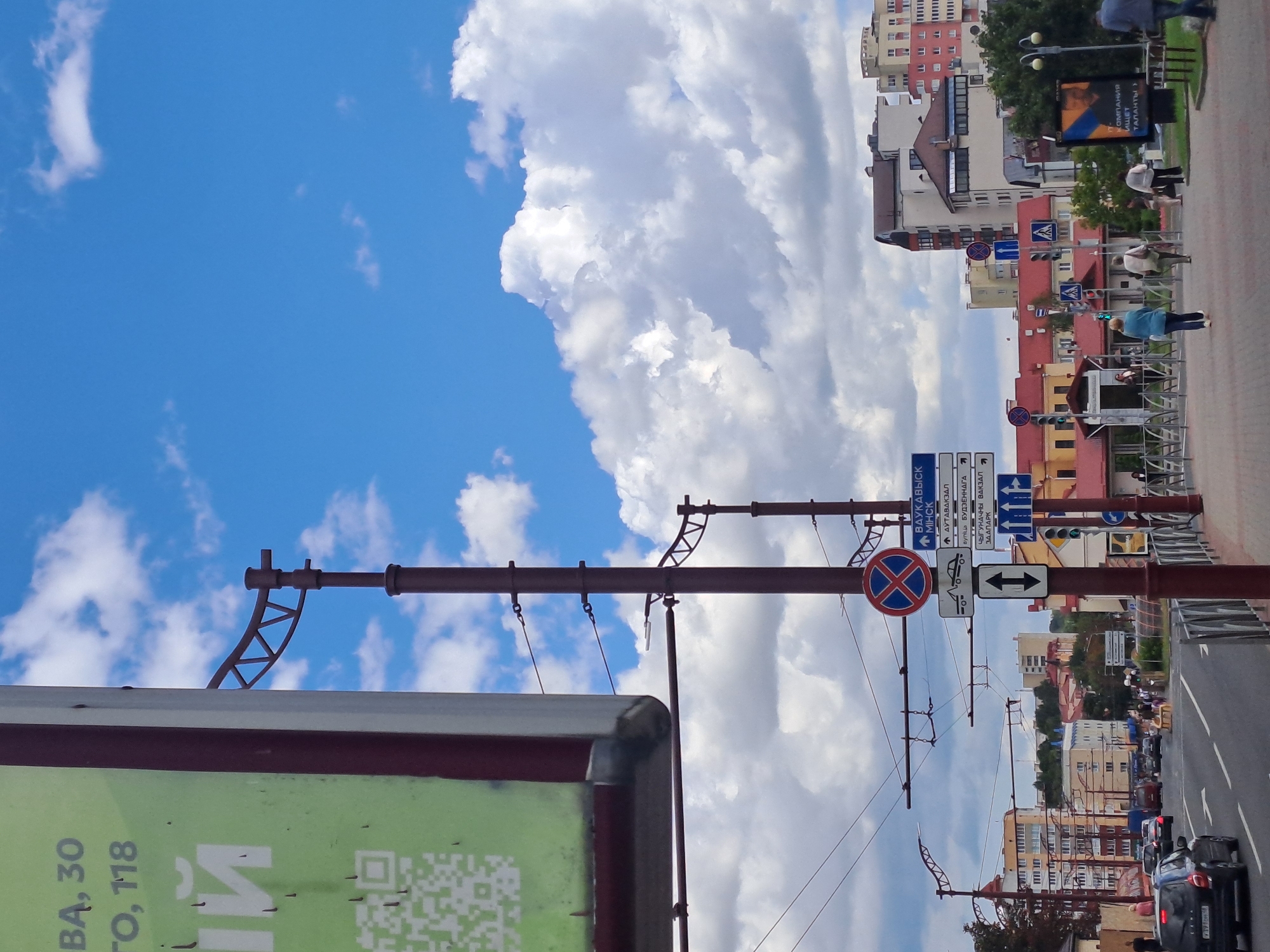 The image captures a street scene in Hrodna, Belarus, on a partly cloudy day. A large green billboard with text and a QR code is visible on the left, partially obscuring a red-brown lamppost with complex wiring. Several street signs are mounted on lampposts, indicating directions and traffic regulations. One sign, with blue lettering, points to "ВарКАШЫСК" (Vaukavysk) and "МIНCK" (Minsk), along with street names. Another sign depicts a tow-away symbol. Traffic lights are visible, showing green. Pedestrians are crossing the street or walking on the sidewalk. Cars are driving on the road, including a black hatchback on the left. Buildings line the street, with a mix of architectural styles. In the background, more buildings and trees are visible under a blue sky with scattered white clouds.