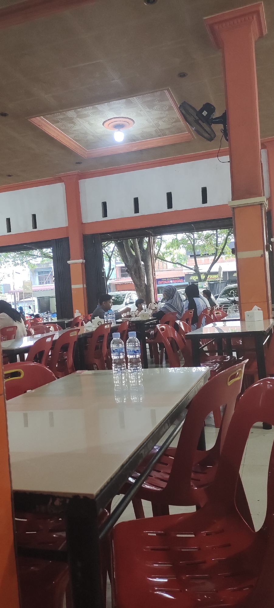 The image depicts the interior of a restaurant in Lambaro, Indonesia. The foreground features a white table with two bottles of water on it. The table is surrounded by red plastic chairs. In the background, several patrons are seated at tables, some of whom are women wearing hijabs. The restaurant appears to have an open-air design, with large windows or openings looking out onto a street with trees and buildings. The ceiling has an orange frame and a patterned panel with a light fixture. A black oscillating fan is mounted on an orange column on the right side of the image. The overall atmosphere suggests a casual dining environment.