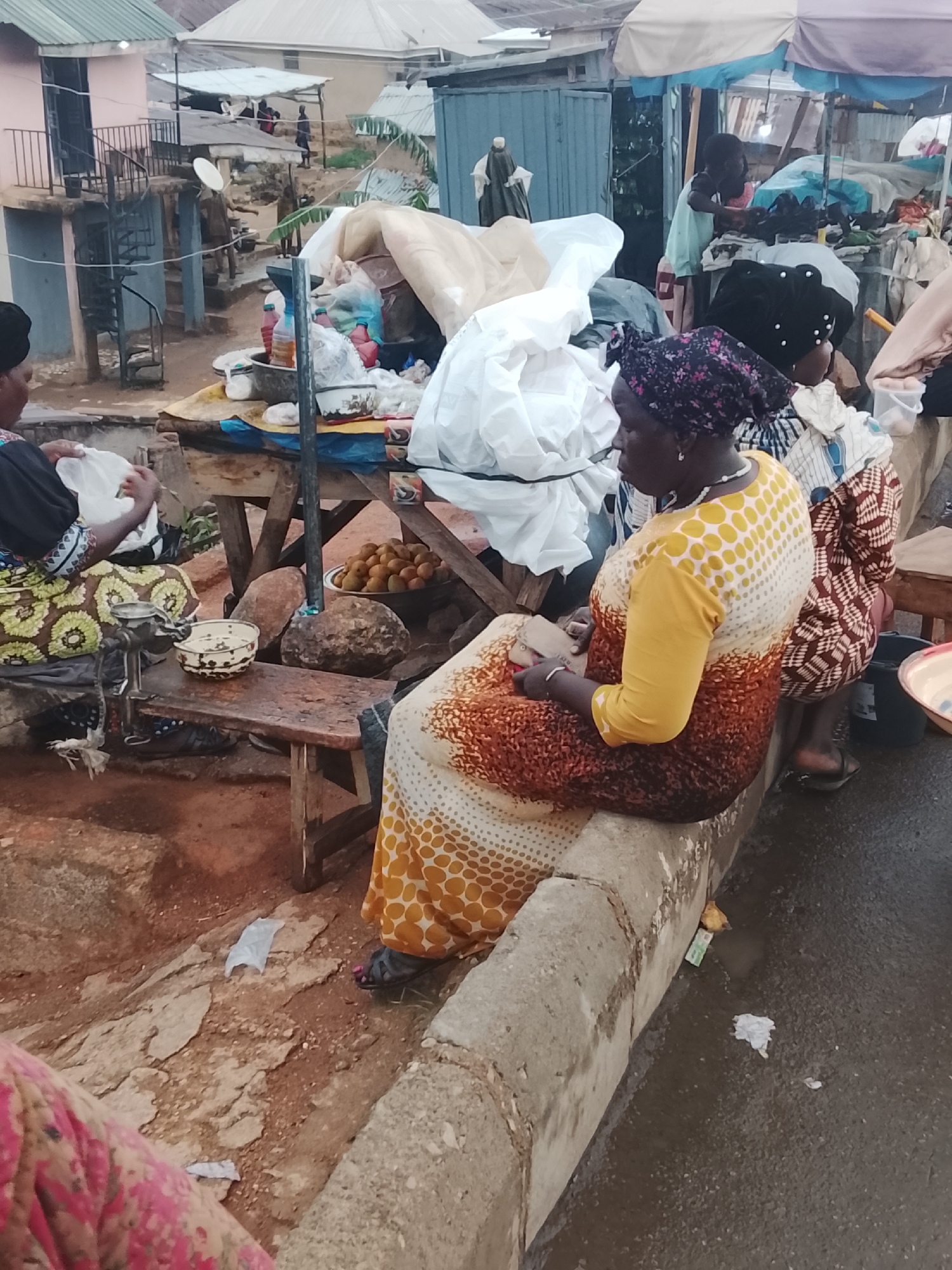 The scene depicts a street market in Gwong, Nigeria. Several women are seated and engaged in commerce. One woman, wearing a yellow and brown patterned dress and a purple headscarf, sits on a concrete curb, holding something in her hands. Another woman, wearing a black top and a green and yellow patterned skirt, sits at a wooden table sorting items into a white plastic bag. On the table are a metal grinder, a white bowl with contents, and a bowl of round fruits. Other women are visible in the background, near stalls with various goods. A metal structure and buildings are visible in the background. The ground is a mix of dirt and pavement.