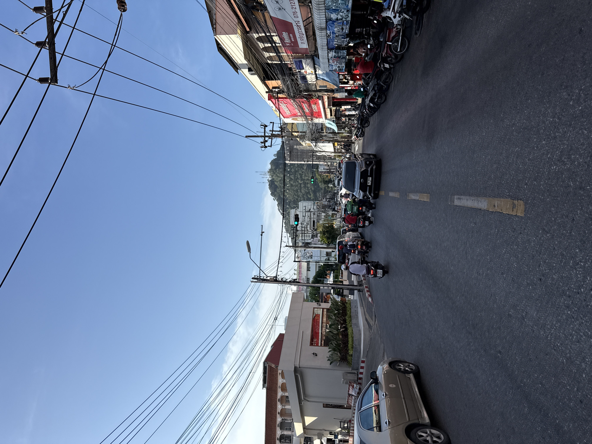 The image depicts a street scene in Phuket City Municipality, Thailand, characterized by a busy road with moderate traffic. A golden sedan is parked on the left side of the road, while a dark SUV is in motion in the center, heading away from the viewer. Multiple motorcycles and scooters are also present, some carrying passengers, navigating the road. The street is lined with buildings on both sides, displaying a mix of architectural styles and commercial signage, some with Thai text. Overhead, a complex network of electrical wires is strung between utility poles, a common sight in many urban areas. In the background, a verdant hill rises, with antennas visible at its peak, under a clear blue sky. The lighting suggests it is either early morning or late afternoon, given the soft sunlight. The overall impression is that of a bustling urban environment in a tropical location.
