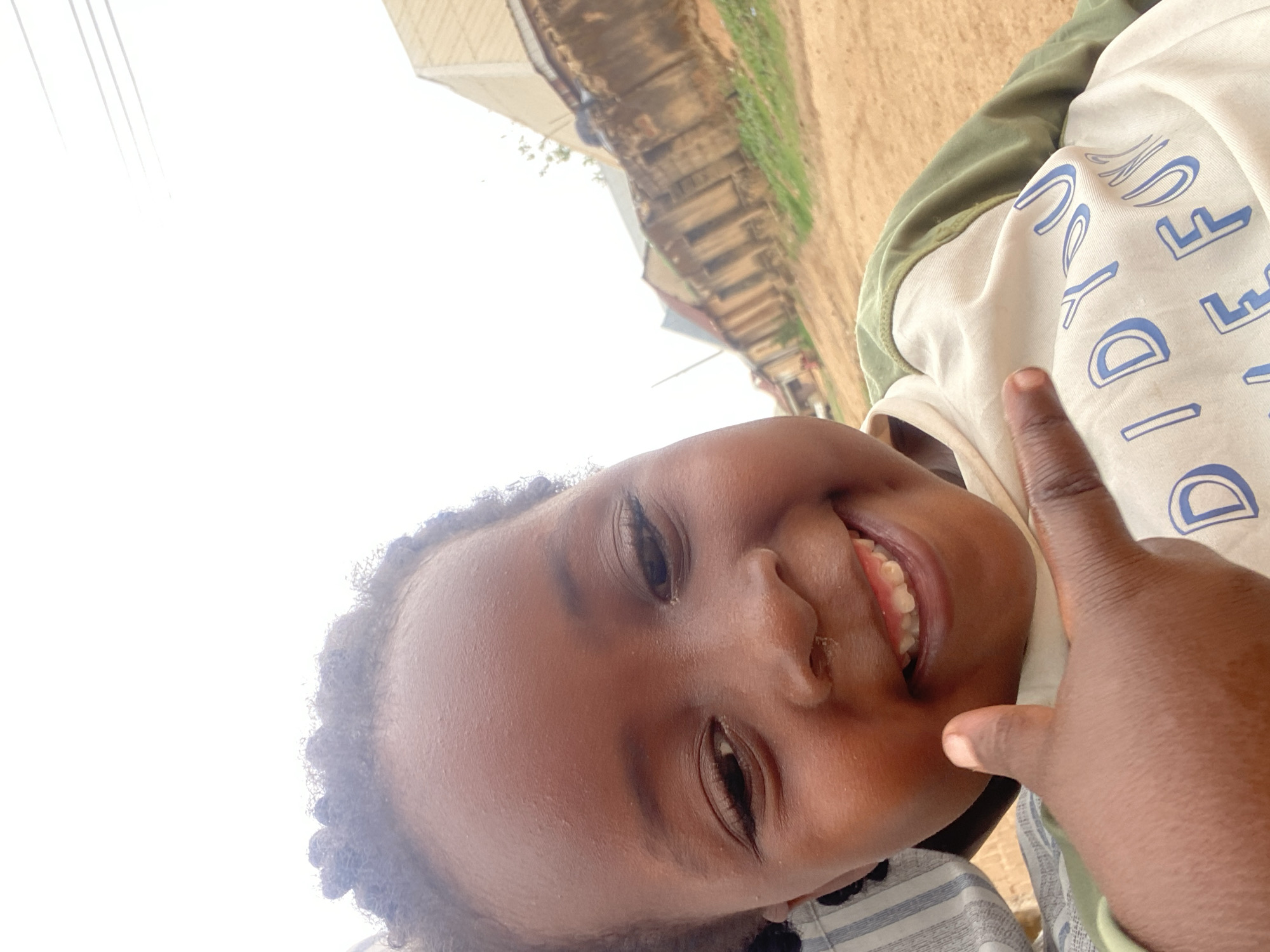 A close-up, tilted shot shows a young child smiling. The child is dark-skinned and wears a light-colored shirt with blue lettering and a green top. A thumb is placed against the child's cheek. In the blurred background, there are structures that appear to be buildings. Based on the location information, this appears to be in an urban or populated environment, specifically Kuru Dungus, Nigeria.