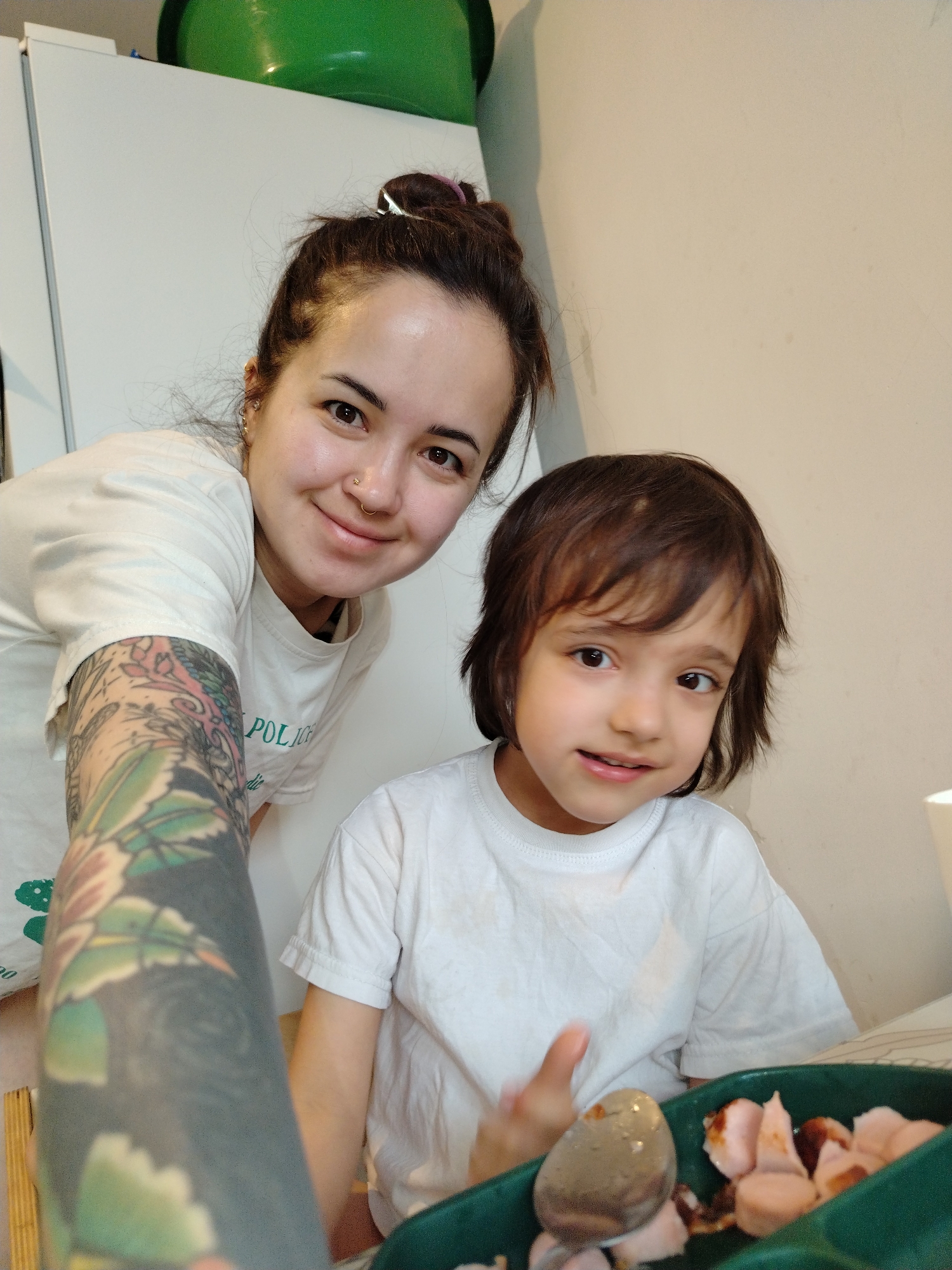 An indoor, eye-level photograph captures a woman and a young child posing together, likely a mother and child. The woman, on the left, leans towards the camera with a gentle smile. She has dark hair styled in a bun, dark eyes, and a nose ring. Her left arm, positioned prominently in the foreground, is adorned with a colorful full-sleeve tattoo featuring intricate floral and leafy patterns. She wears a white t-shirt with a partial green graphic and text, where the word "POLICE" or similar is partly visible.
Beside and slightly below her, on the right, is a young child with shaggy brown hair and wide brown eyes, also looking at the camera with a soft smile. The child is wearing a plain white t-shirt. The child's right hand is raised near a green, compartmentalized tray in the lower right corner of the frame. The tray contains cut-up food resembling sausages or hot dogs, alongside a spoon holding another food item.
The background shows a white cabinet or refrigerator on the left, with a green bowl or container placed on top. The wall behind the child is a plain, light color. The scene suggests a relaxed, happy moment in a home setting, possibly a kitchen or dining area, during daytime. The location context is Noviy, Russia.