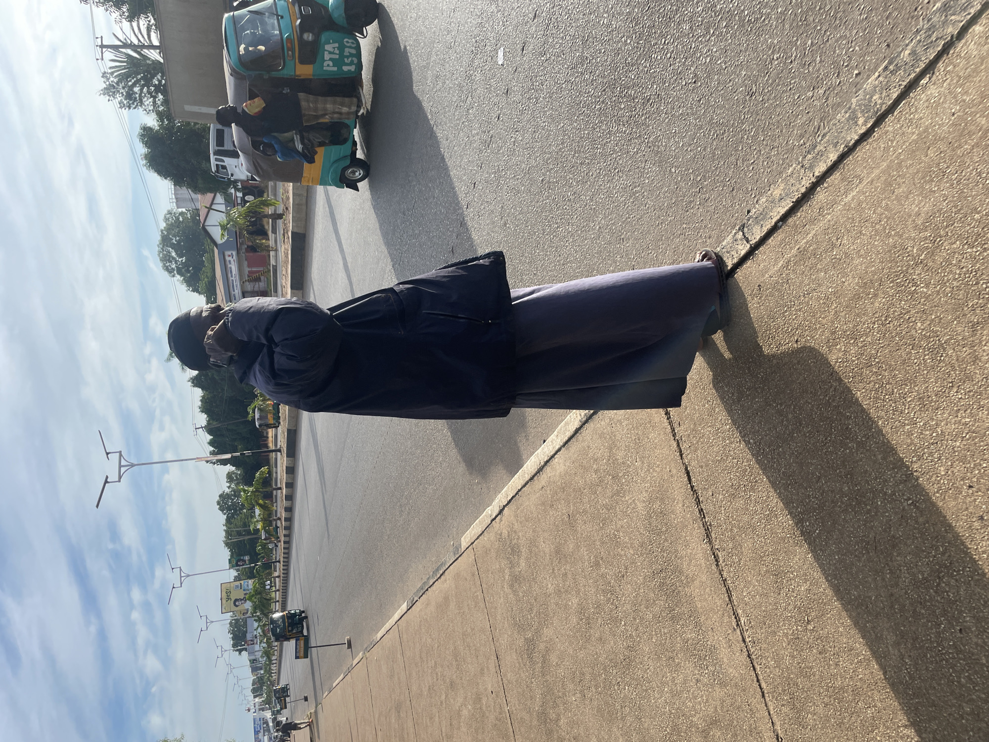 In Jos, Nigeria, a man wearing a dark blue jacket and a long, gray, pleated skirt stands on a paved sidewalk, talking on a mobile phone. His shadow is cast across the sidewalk and onto the adjacent asphalt road. A blue and yellow motorized tricycle, with "PTA 1578" visible on its side, is driving on the road. Two people are visible within the tricycle, one of whom appears to be the driver. The road is lined with palm trees, streetlights, and buildings. The sky is partly cloudy.