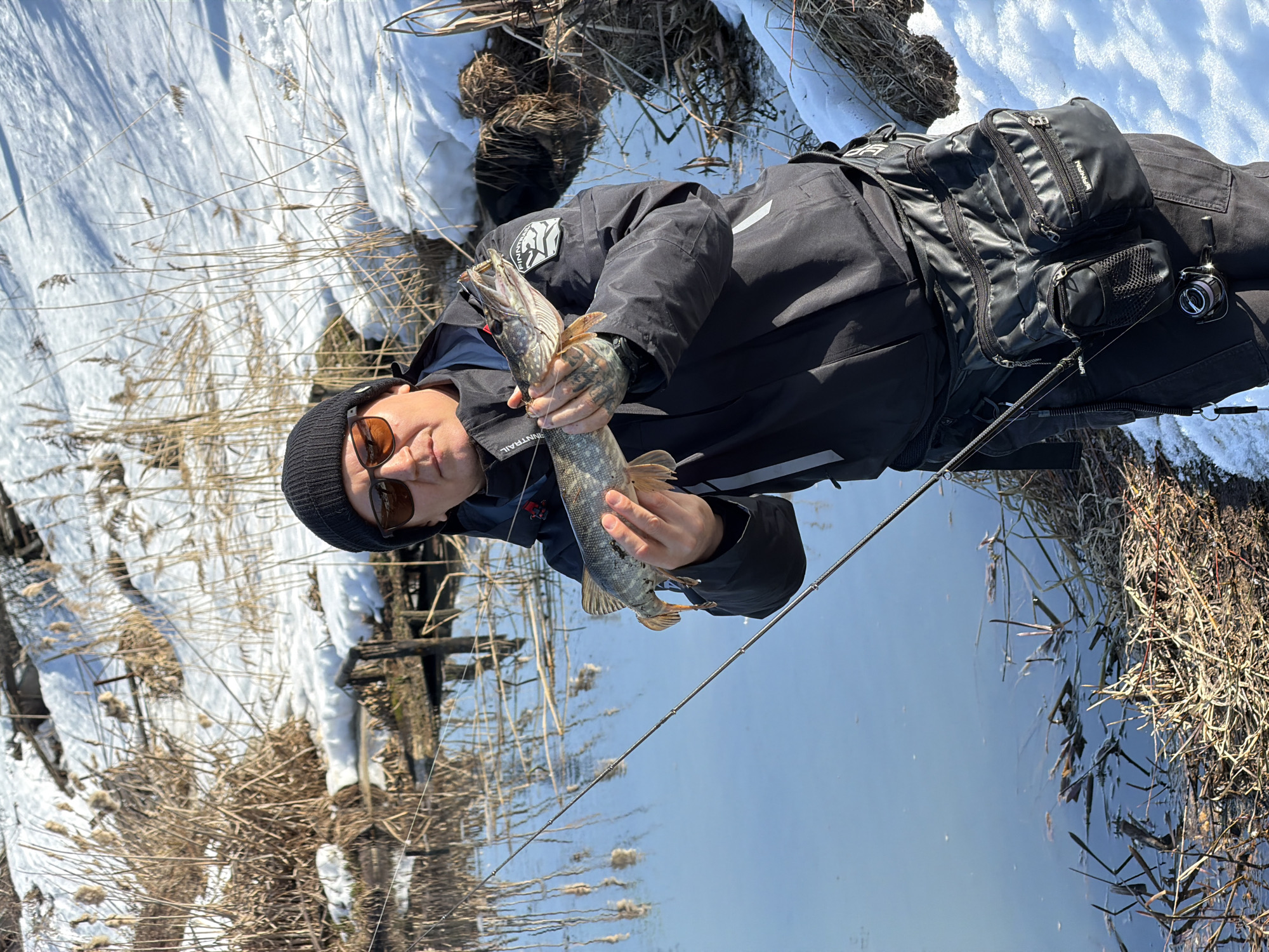 A man stands on a snowy bank next to a body of water, holding a Northern Pike. He is wearing a black beanie, sunglasses, a black jacket with a white stripe and the word "FINNTRAIL" on the sleeve, and a black chest pack. He has tattoos on his hands. The surrounding environment includes snow, reeds, and what appears to be a river or lake. Based on the image metadata, the location is likely Chaikovsky, Russia.