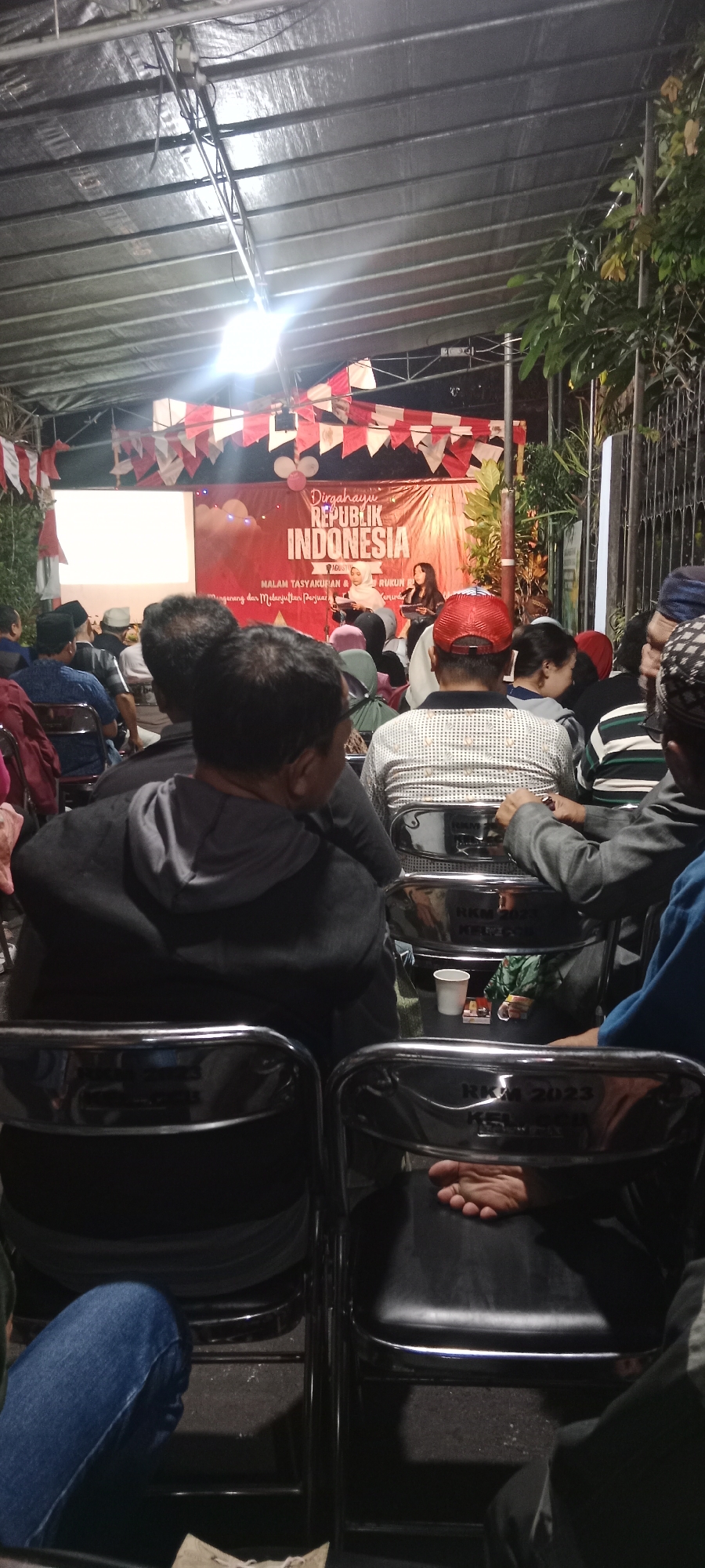 The image depicts an outdoor event at night, possibly a community gathering or celebration, set in Magelang, Indonesia. The scene is illuminated by artificial lighting, casting shadows and highlighting the assembled crowd.
In the foreground, several individuals are seated on chairs, facing a raised platform or stage. Their backs are mostly turned to the camera, and they are dressed in casual attire, with some wearing head coverings. The chairs themselves are simple, dark-colored with chrome-plated frames, some bearing engraved text, including "RKM 2023".
On the stage, a banner with red and white triangular pennants is visible. The banner features text that appears to be in Indonesian, with prominent lettering "Dirgahayu REPUBLIK INDONESIA" (Long Live the Republic of Indonesia). Two women are standing on the stage, one in a white hijab and the other in dark clothing, seemingly addressing the audience or participating in a program. There is a screen or projector visible to the left of the stage, displaying a blank white area.
The environment suggests a temporary setup for an event, with a large canopy or roof structure overhead, supported by metal framing. Green foliage is visible to the right of the stage, indicating an outdoor setting with some natural elements. The overall impression is of a local event attended by members of the community in Magelang.