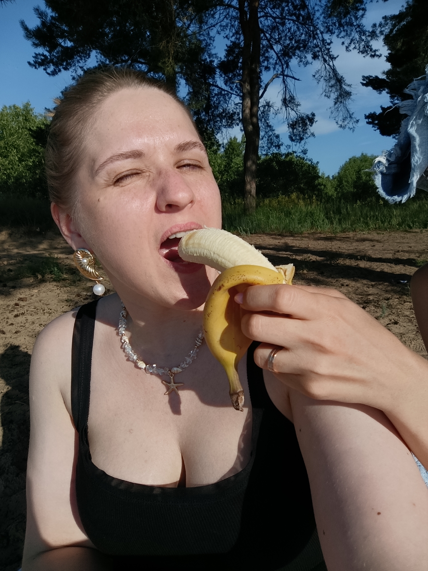 A woman is sitting outdoors in a sandy area with trees and green foliage in the background. She is wearing a black tank top and a multi-strand necklace with a starfish pendant. She has a large, ornate earring on her left ear. A partially peeled banana is in her right hand, and she is eating it. Her eyes are closed, and her mouth is open around the banana. A ring is visible on her right ring finger. The sky is blue with some clouds.