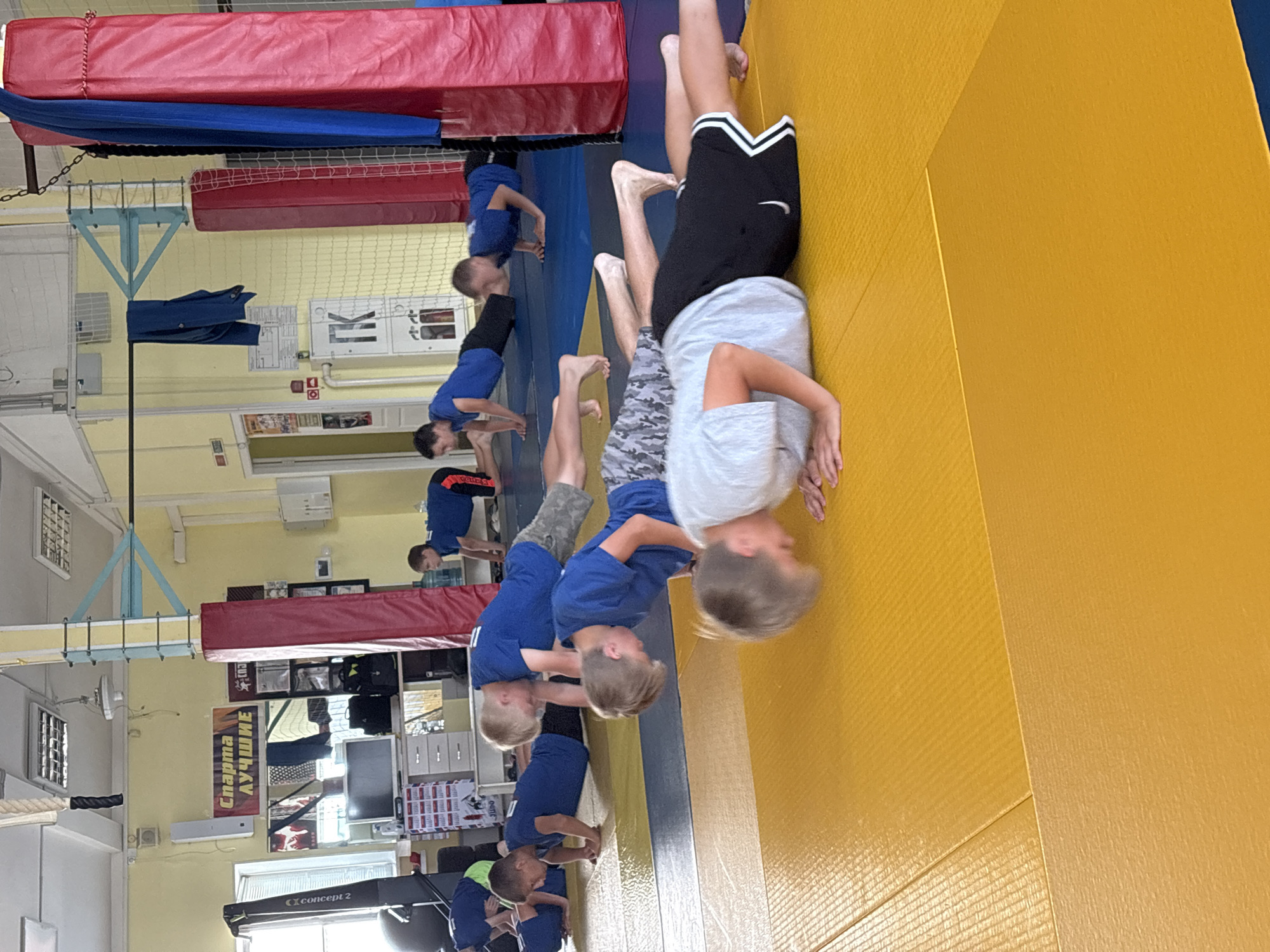 The image depicts a group of young boys performing push-ups on yellow and blue mats in what appears to be a gymnasium. The boys are wearing athletic clothing, predominantly blue t-shirts and shorts. In the background, there are training equipment, banners with Russian text, and gym equipment like a rowing machine and a boxing ring post. The overall environment suggests a sports training facility. The banner in the background reads "Спарта Лучшие" which translates to "Sparta Best". Based on the text on the banner and the gym equipment, the location is likely a sports club or training center in Chaikovsky, Russia.