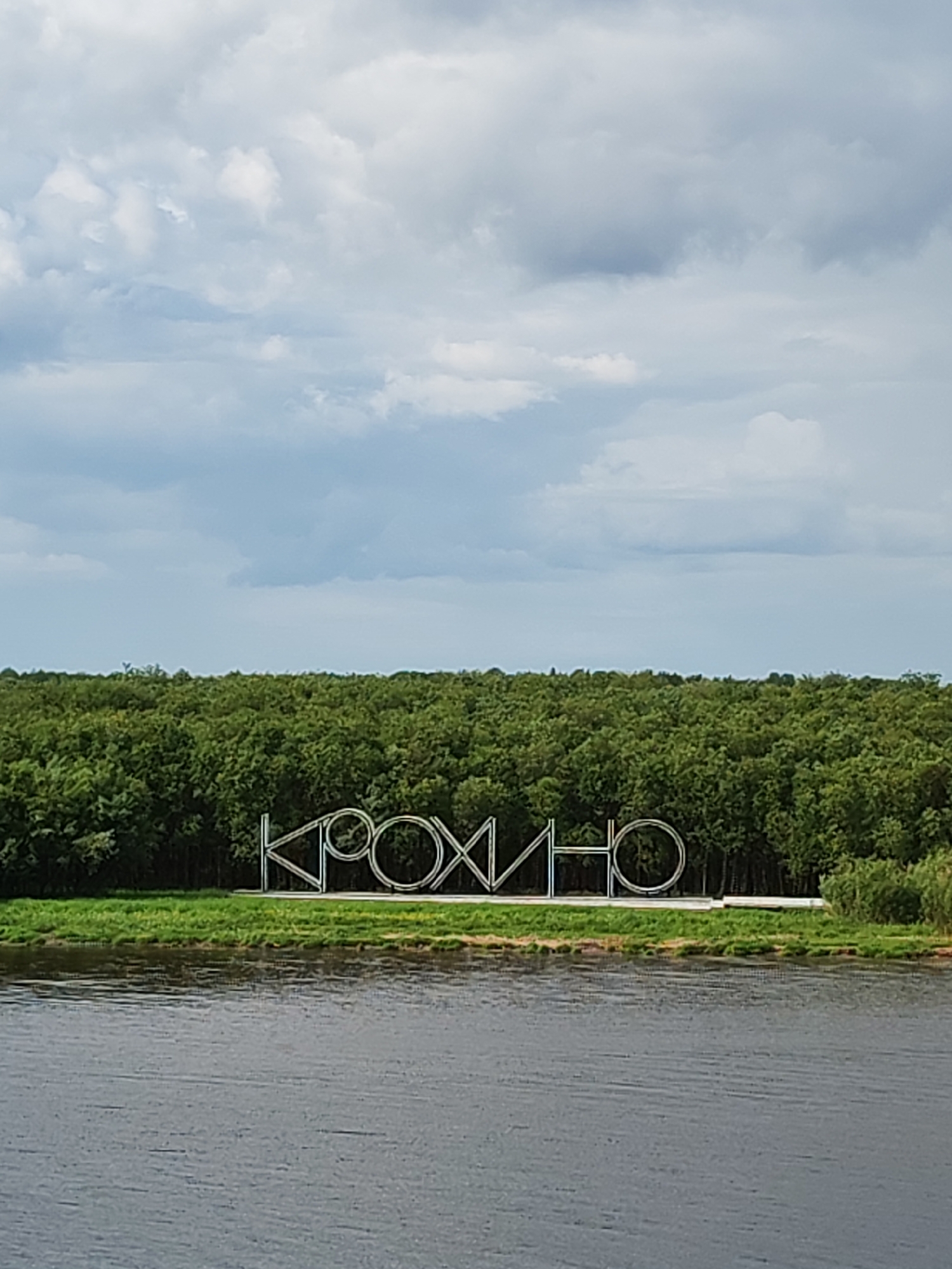 The image shows a view of a body of water, likely a river, with a lush green forest visible on the opposite bank. In front of the forest, on a strip of grass, is a large, silver metallic sign that spells out "КРОХИНО", which is the name of the location in the city of Krokhino, Russia. The sky is partly cloudy with a mix of gray and white clouds. The water surface appears calm.