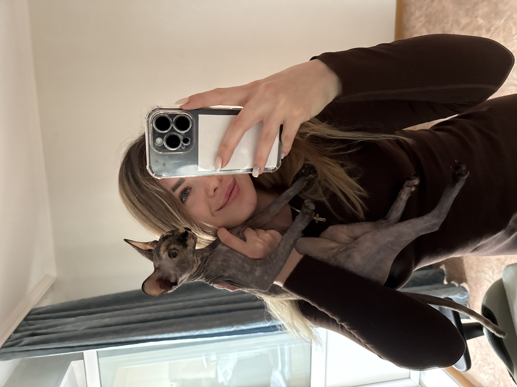 A young woman is holding a hairless cat and taking a selfie with her smartphone. The woman has light blonde hair and is wearing a brown, long-sleeved garment. The cat, gray with dark patches, is held in her arms. The phone has a clear case and a white square obscuring something on the back. The background includes a window with gray curtains and a glimpse of what appears to be furniture. The location appears to be an interior space. The specific city is not ascertainable from the image.