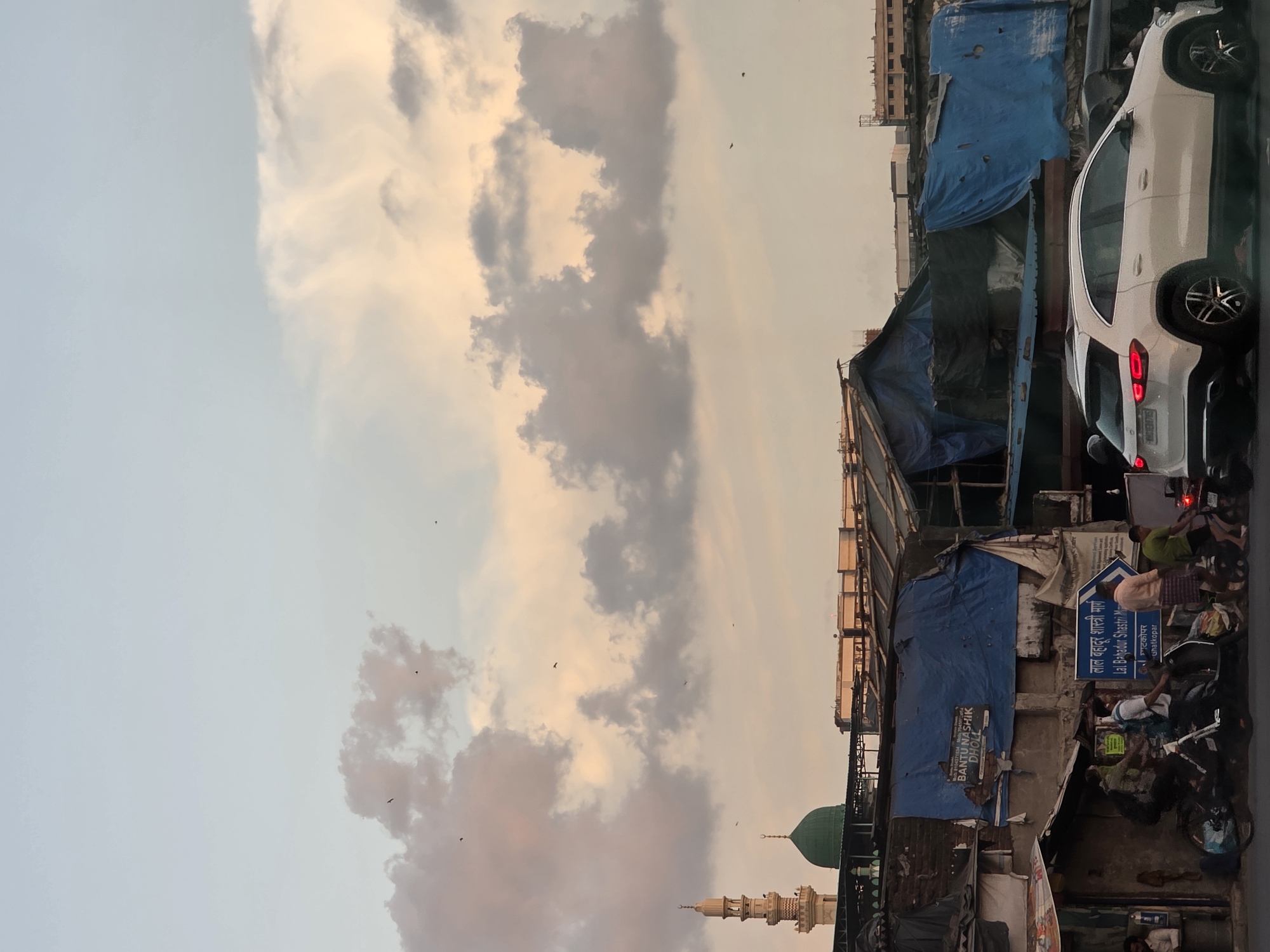 The image depicts a street scene in Mumbai, India, likely during late afternoon or early evening, given the soft lighting and pastel hues of the sky and clouds. In the background, a mosque with a minaret and green dome stands out against the sky. Below it, makeshift structures covered with blue tarpaulins suggest a densely populated urban area, possibly a slum or informal settlement.
In the foreground, a white SUV is parked on the street, its brake lights illuminated. Several people are visible in the mid-ground and foreground. Some are on motorcycles, while others are walking. A street sign with blue lettering and an arrow points to "LAL BAHADUR SHASTRI MARG". The overall impression is of a vibrant, albeit somewhat chaotic, urban environment characteristic of many parts of Mumbai.