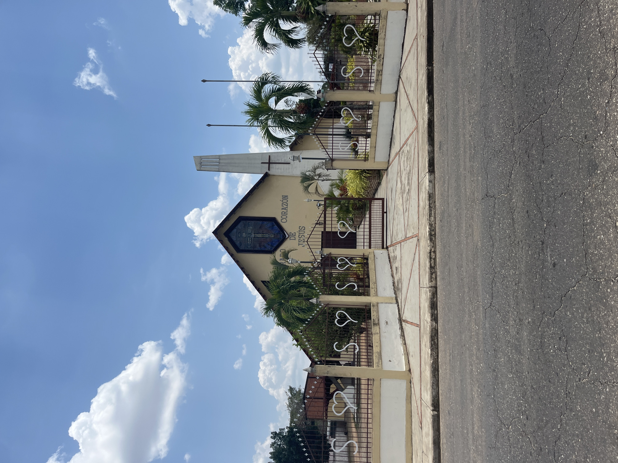 The image depicts the Sacred Heart of Jesus Church (Iglesia Sagrado Corazón de Jesús) in Tinaquillo, Venezuela. The church is a beige-colored building with a pointed roof, featuring a stained glass window with a cross. Its name is written vertically on the facade. A tall, rectangular tower stands adjacent to the main structure. A decorative metal fence encloses the property, adorned with heart-shaped elements. Palm trees are visible within the church grounds. The sky is blue with scattered white clouds. A section of sidewalk and a paved road are in the foreground.