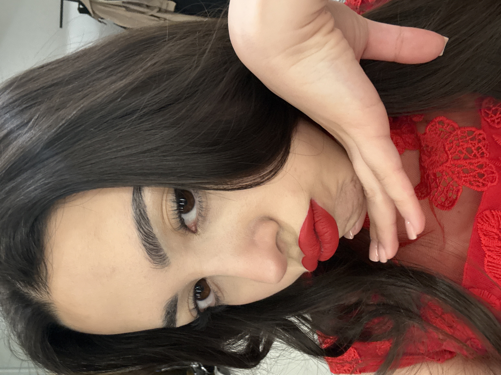 A woman with dark hair and brown eyes is lying down, wearing red lipstick and a red, lacy garment. She is positioned at an angle to the camera, with her hand partially covering her face. The background is indistinct, showing only glimpses of furniture and a light-colored wall. No information suggests this photo was taken in Yerevan, Armenia.
