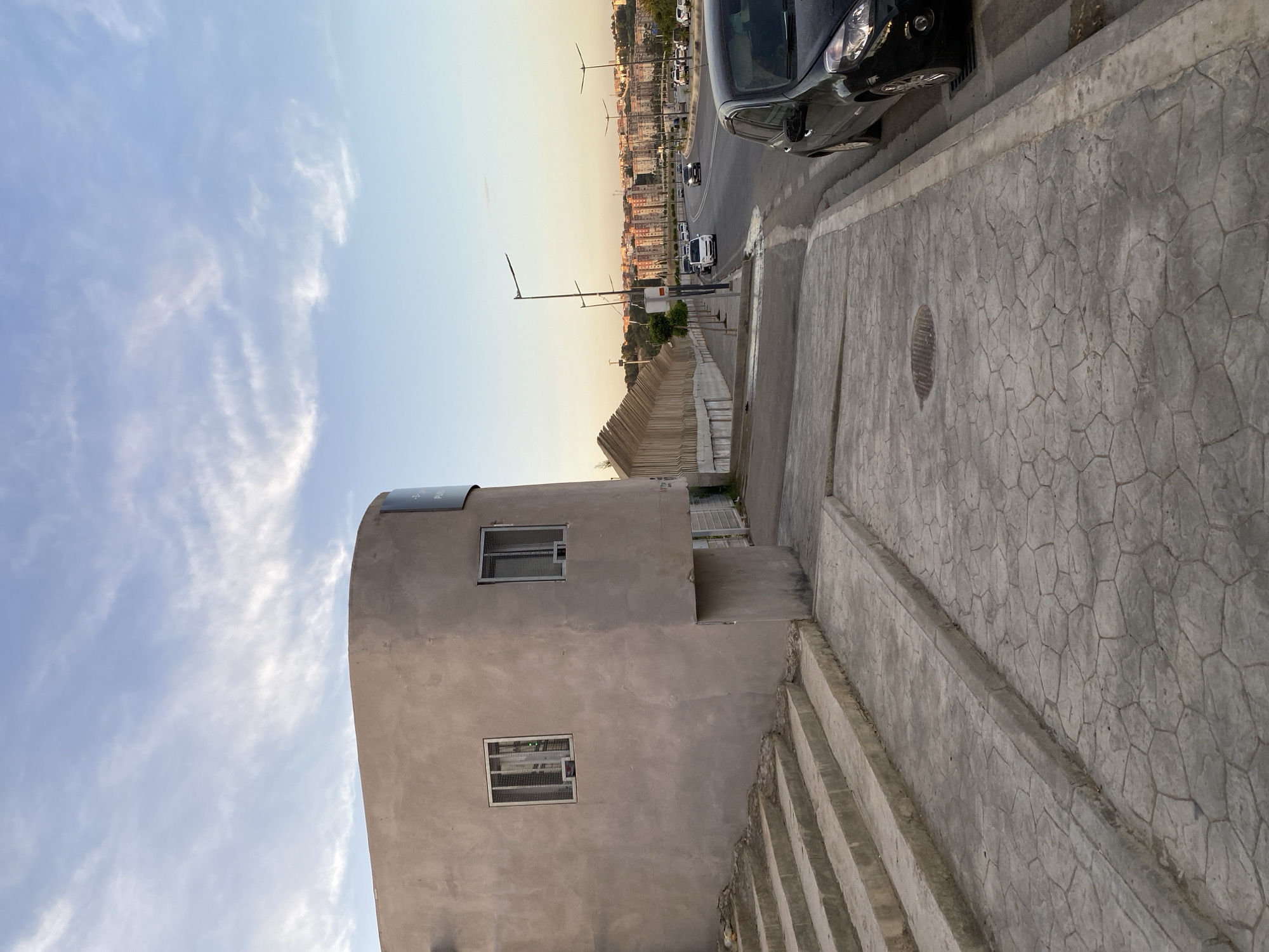 The image captures a street scene in Douera, Algeria, during the late afternoon. In the foreground, a textured concrete sidewalk with a hexagonal pattern leads to a set of concrete stairs. To the left, a modern building with a curved facade, constructed from light brown plaster, features two windows with metal grates. Adjacent to the building, a dark-colored car is parked on the right side of a paved road. The road curves to the right and continues into the distance, lined with streetlights. Beyond the road, a long, segmented wall stretches into the background. In the far distance, a densely populated urban landscape with apartment buildings is visible, set against a backdrop of hills under a sky with scattered clouds. The overall lighting suggests the golden hour before sunset.