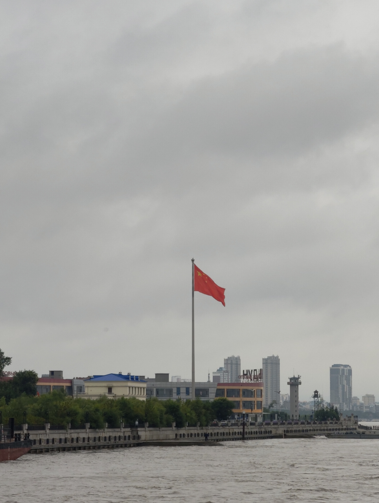 The image captures a cloudy day scene overlooking a body of water, likely a river, with a city skyline in the background. A prominent Chinese flag flies on a flagpole in the mid-ground. The shoreline features a paved walkway with decorative railings, bordered by lush green trees and several buildings. Among these structures is a distinctive building with a blue roof. Further into the cityscape, various modern buildings, including high-rises and a lighthouse-like tower, are visible. The water is choppy, suggesting a breeze. A barge or ship is partially visible on the left side of the frame. The overall impression is of an urban waterfront on an overcast day. While the prompt mentions 幸福乡, China, there are no specific visual cues within the image that definitively identify this particular location.