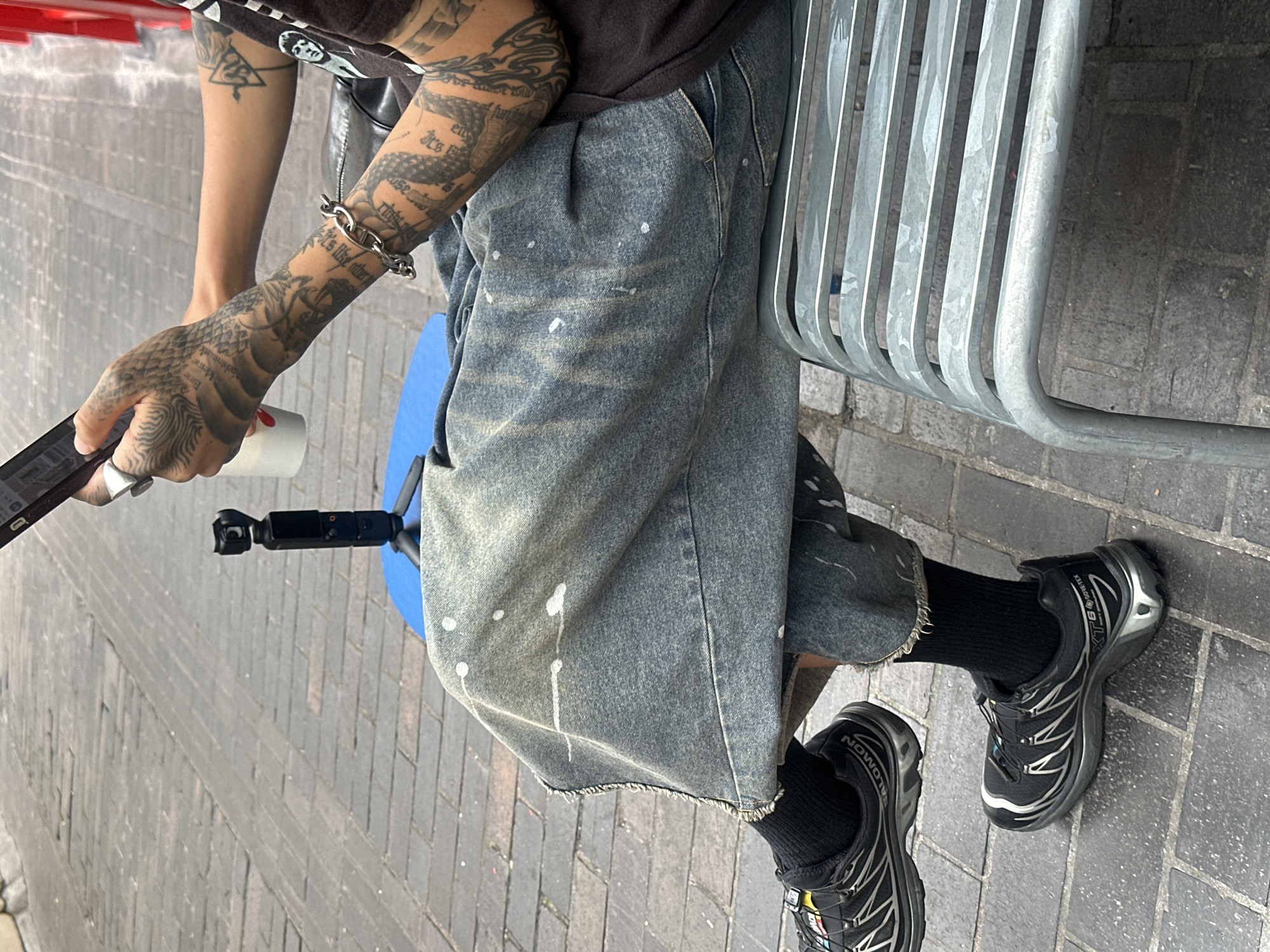 The image shows a person sitting on a metal bench on a brick-paved street. The person is wearing wide-leg denim pants with paint splatters and black Salomon XT-6 Gore-Tex sneakers. They are holding a smartphone and a white cup. A small black gimbal is set up on a blue mat to the left of the person. The person has extensive tattoos on their arms and hands. The environment suggests an urban setting, likely a street or public area. The bricks are laid in a basketweave pattern. The lighting indicates it is daytime, with shadows visible on the pavement. The background is blurred, but appears to be a street with more brickwork and some red elements. The overall impression is a candid moment of someone engaged with their phone and camera equipment in a public urban space.