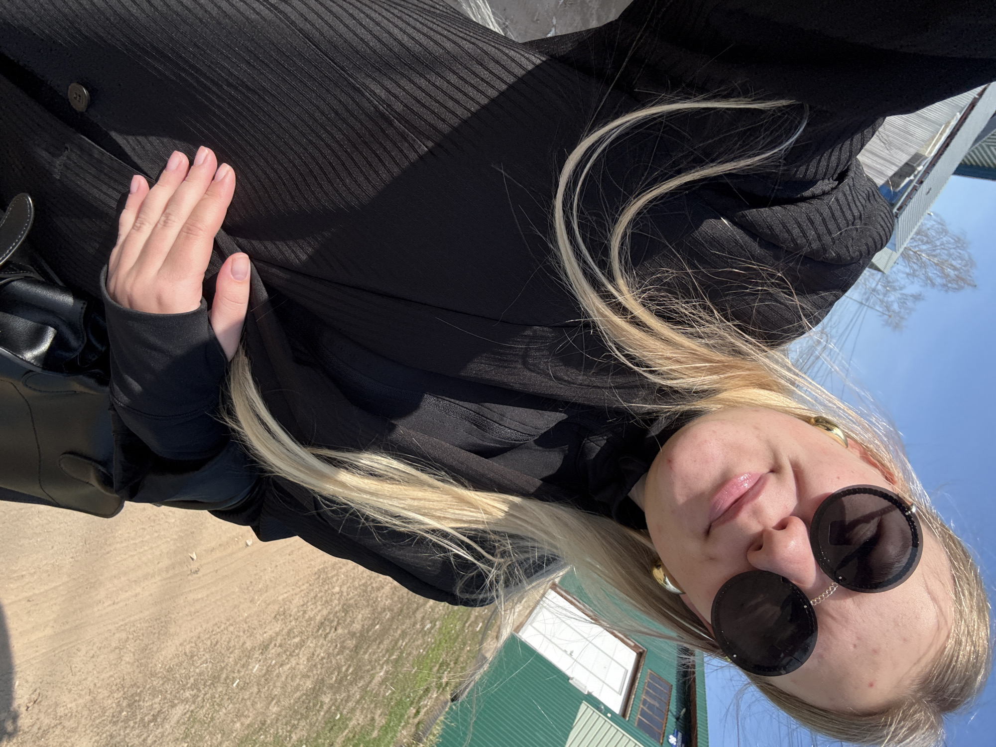 The image, oriented correctly, depicts a young woman outdoors under a clear, bright blue sky. She is seen from the chest up, looking upward towards the camera with a neutral to slightly smiling expression, her lips slightly parted and glossy. She has long, blonde hair that flows over her shoulders and across her black attire.
She is wearing large, round, dark sunglasses connected by a thin chain across the bridge of her nose, obscuring her eyes. Gold-colored hoop earrings are visible on both ears. Her clothing consists of a black, ribbed, long-sleeved garment, possibly a dress or top, made of a textured fabric. Her left hand, with neatly manicured nails and light-colored polish, is gently placed on her stomach area. A black bag or purse is visible on her left side.
The background shows an outdoor setting during daytime with strong sunlight casting shadows. To her left, there is light brown, sandy ground with sparse vegetation. To her right, a building with vibrant green corrugated metal siding and white-framed windows stands under the blue sky. A tree with bare branches is visible in the distance against the sky. This scene is located in Chaikovsky, Russia. No visible text is present in the image.
