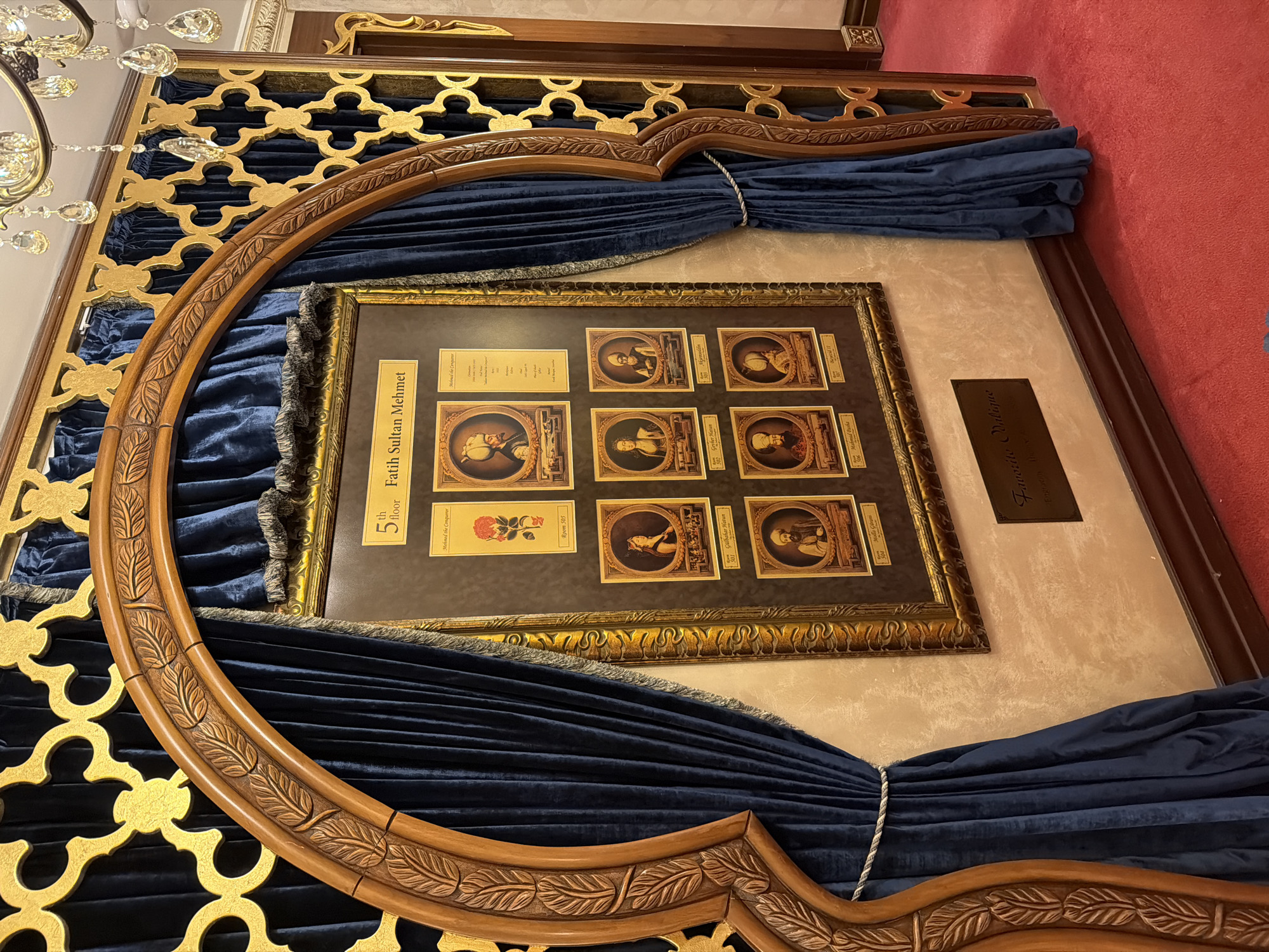 This image depicts a framed display commemorating "Fatih Sultan Mehmet" on the 5th floor. The display features a central portrait of a rose and several smaller portraits of individuals, each with a label and a room number. The frame is ornate, with dark blue velvet curtains draped to the sides, held back by decorative cords. The entire display is set within a larger architectural feature, possibly an archway, with carved wooden elements and a gold filigree pattern on the wall behind the curtains. Below the framed display, a plaque reads "Favorito Valide." The floor is covered in a vibrant red carpet, and a crystal chandelier is visible in the upper right corner. The scene appears to be an interior setting, likely in a historical or cultural establishment. Given the subject matter and the context suggested by the opulent decor, this display is likely located within the Dolmabahçe Palace in Beyoğlu, Istanbul, Turkey, a significant historical landmark.