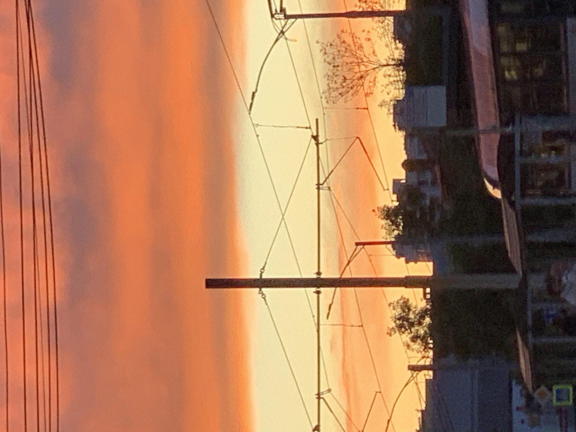 The image displays an urban or suburban landscape captured during either sunrise or sunset, evidenced by the vibrant, warm colors dominating the sky. The photograph is oriented vertically.
On the left side of the image, a striking gradient of colors transitions from a deep, reddish-orange on the far left to a brighter, soft yellow-orange towards the right, resembling a dramatic sky. Faint, horizontal cloud patterns are visible within this colorful expanse.
Across the mid-section and right-middle of the image, an extensive network of dark, silhouetted infrastructure is prominent. This includes multiple thin vertical utility poles, some with horizontal crossbeams, and numerous overhead electrical wires stretching diagonally and vertically. This setup is characteristic of power lines or a catenary system for electric public transport, such as trams or trolleybuses. On the far left, several parallel, thin vertical lines are visible, likely additional overhead wires.
The rightmost portion of the image features dark silhouettes of buildings and trees, providing a clear horizon line against the luminous sky. Scattered trees, some with bare branches, are discernible, particularly in the upper right section. The outlines of various buildings are visible along the right edge, with some faint rectangular shapes indicating windows within the darker structures. A large, dark, elongated shape, possibly part of a vehicle like a tram or train, is partially visible along the far-right edge, though its details are obscured.
In the bottom-right corner, a small, bright green square with a yellow interior and a dark dot or symbol is present, resembling a sign, but no legible text is visible. The scene conveys a tranquil and atmospheric moment as day transitions to night or vice versa.