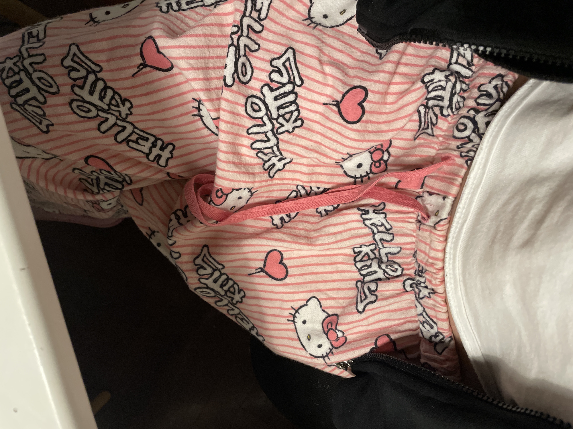 The image shows a close-up of pink striped pajama pants with a Hello Kitty pattern. The pants have a pink drawstring and are positioned next to a folded white garment, a black garment, and a white cupboard.