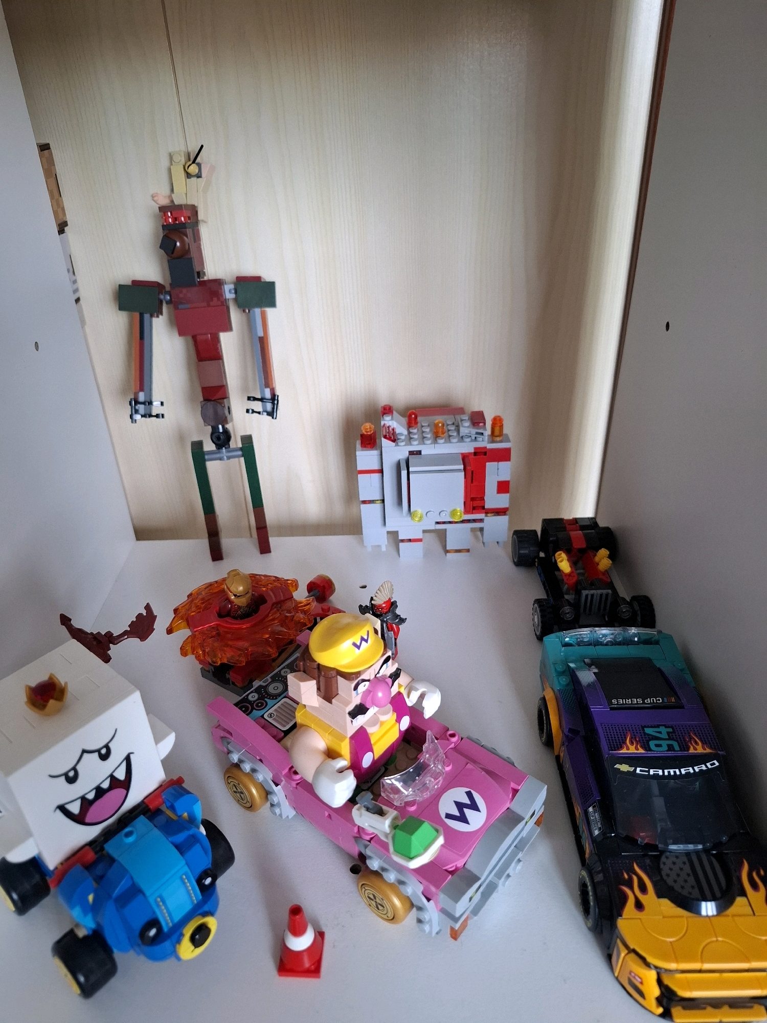 The image shows a collection of LEGO toys on a shelf. On the left, there is a white LEGO figure resembling a ghost with a crown and a blue vehicle. Next to it is a pink LEGO kart with a figure resembling Wario. To the right of the kart is a black and teal LEGO car with flame decals and "Camaro" written on it. In the background, there is a tall, red and gray LEGO robot and a white and red LEGO structure resembling a building. The shelf appears to be made of light-colored wood. The image was taken in Tyumen, Russia.
