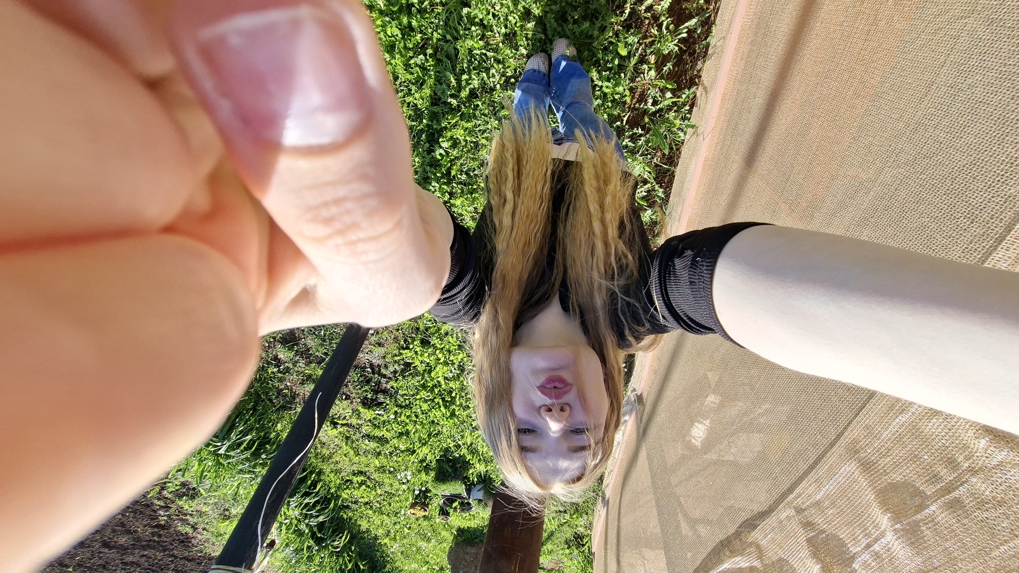 A female subject is seen in a self-portrait photograph, upside down relative to the viewer's perspective. The camera angle is low, looking up. She is wearing jeans and shoes, with her blond hair hanging down towards the grass. A part of her face, including her nose and mouth, is visible. One of her hands is prominent in the foreground, with a visible fingernail. The environment includes grass, a brown fabric-like structure, and what appears to be a pole or pipe, suggesting an outdoor setting in Большой Букор, Russia.