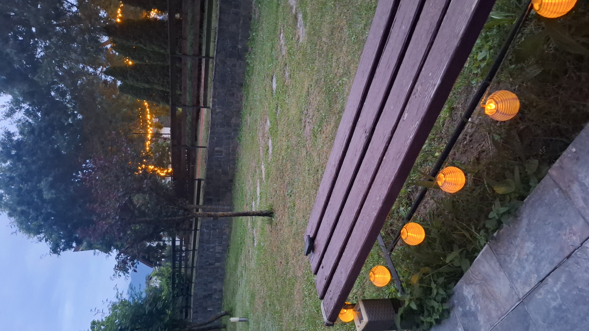 This media file depicts an outdoor scene at dusk in Razlog, Bulgaria. The main subject is a wooden bench, positioned at an angle in the foreground, with its textured surface and deep purple hue clearly visible. Along the edge of the bench, a string of small, orange, spherical lanterns are illuminated, casting a warm glow on the surrounding grass and soil. These lanterns are spaced evenly and add a festive or decorative element to the setting.
Behind the bench, a grassy area stretches out, punctuated by a few scattered patches of darker soil. A stone wall forms a boundary further back, and beyond that, trees with dark green foliage are visible. In the distance, more strings of warm yellow lights are draped across trees, creating a magical atmosphere as daylight fades. The sky above is a deep, muted blue, indicating the time of day is either early evening or twilight.
There are no people or animals visible in the frame. The setting appears to be a garden or a park area, possibly in a rural or semi-rural location given the natural elements and the rustic appearance of the bench and stone wall. The weather seems calm, with no indication of wind or rain. The overall mood conveyed is peaceful and inviting, enhanced by the soft lighting and the natural surroundings. No text is visible in the image.