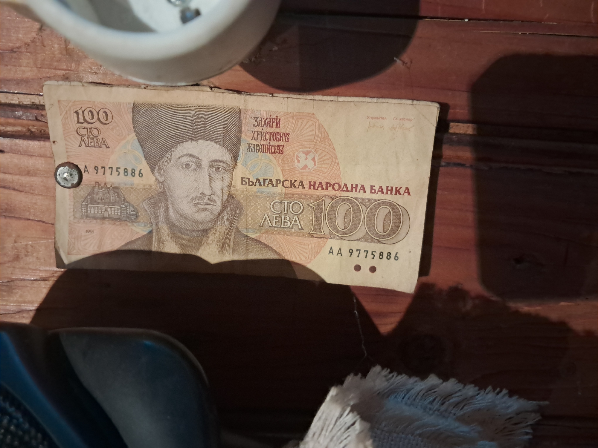 A close-up shot shows a 100 Bulgarian lev banknote from 1991 lying on a wooden surface. The bill features a portrait of Zahari Hristovich Zograf, a Bulgarian painter, along with the text "БЪЛГАРСКА НАРОДНА БАНКА" (Bulgarian National Bank) and the value "100 СТО ЛЕВА". The serial number "A 9775886" is printed on the left side of the note, and "AA 9775886" on the right. A metal tack pierces the upper left corner of the bill. A white ceramic object, possibly a cup, is partially visible at the top of the frame, casting a shadow on the banknote. Another object is partially visible in the lower left corner, and white fabric is partially visible in the lower right corner. There is no indication of Seykovtsi, Bulgaria.