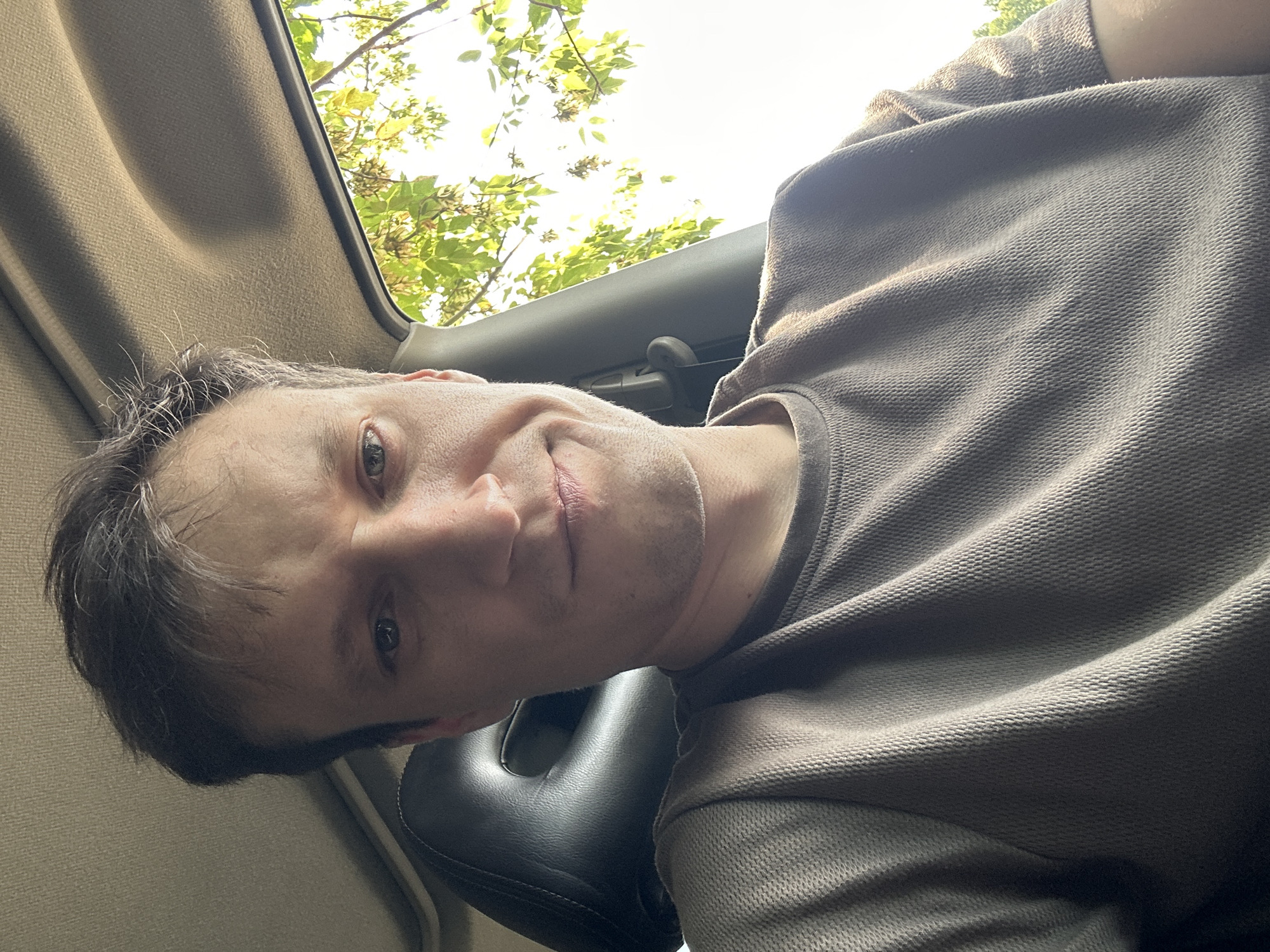 A close-up, waist-up, selfie taken from inside a car shows a fair-skinned man with short, dark brown hair and blue eyes. He is looking directly at the camera with a slight smile. He is wearing a textured, taupe t-shirt. The man's head is angled slightly to the left, with his left eye and cheek more visible than his right. The interior of the car is visible, including a dark gray headrest behind him and the car's headliner above. Sunlight filters through the leaves of a tree outside the car window on the right side of the frame. The man is positioned in the driver's seat, suggesting a location within the city of Makiv, Ukraine, given the context.