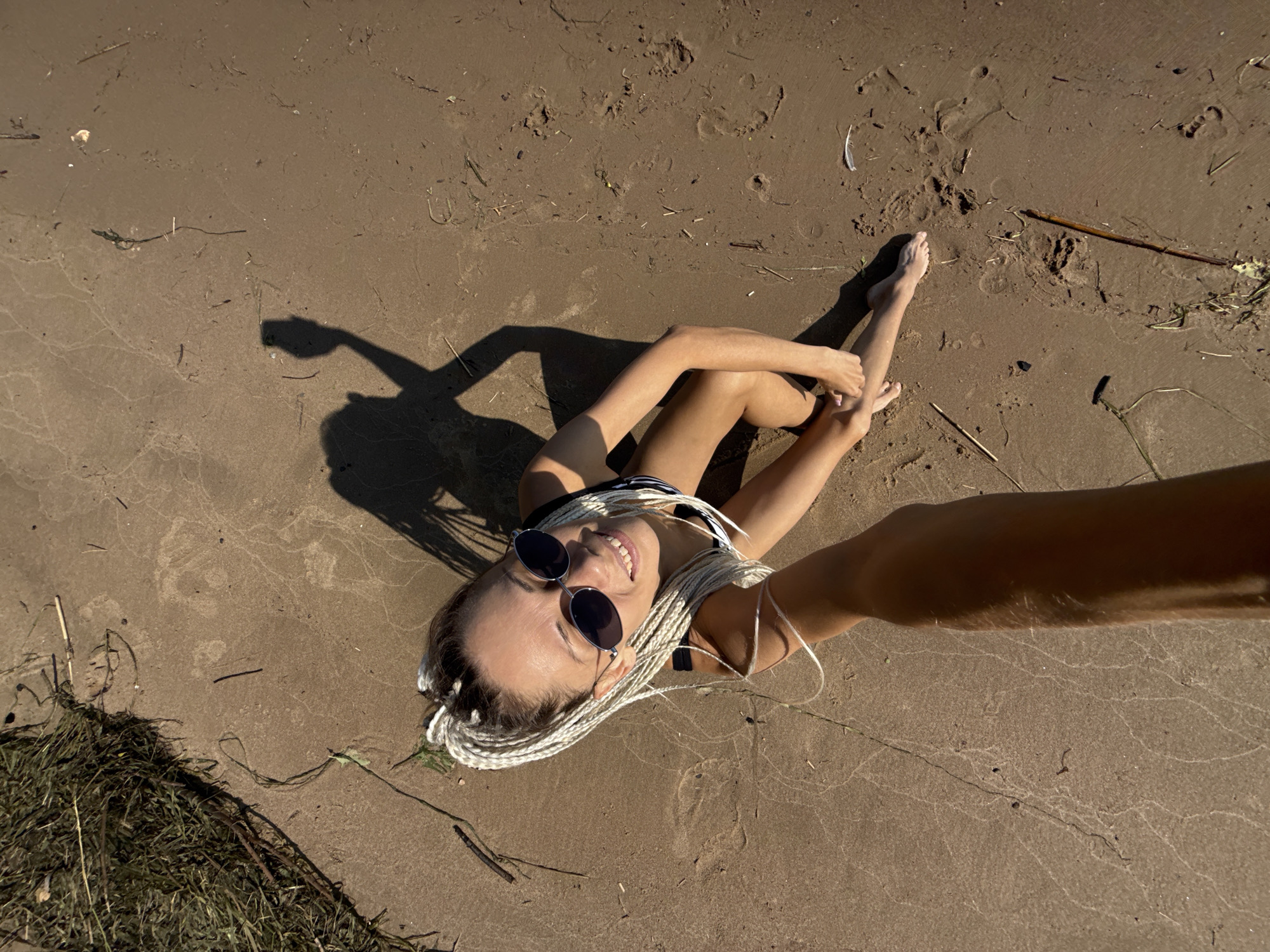 A top-down, full shot shows a woman with blonde braided hair and sunglasses sitting on a sandy beach, facing upwards and smiling at the camera. Her left arm is extended towards the camera, holding a phone out of view, creating a selfie. She is wearing a black and white striped swimsuit. The sand is light brown and textured with footprints and scattered debris, including small sticks and dry grass. In the upper left corner, a patch of green grass and vegetation is visible. The shadow of the woman and the phone she is holding is cast on the sand to her right. The scene appears to be outdoors, with natural lighting. The location is identified as a beach in the city of Dumino, Russia.